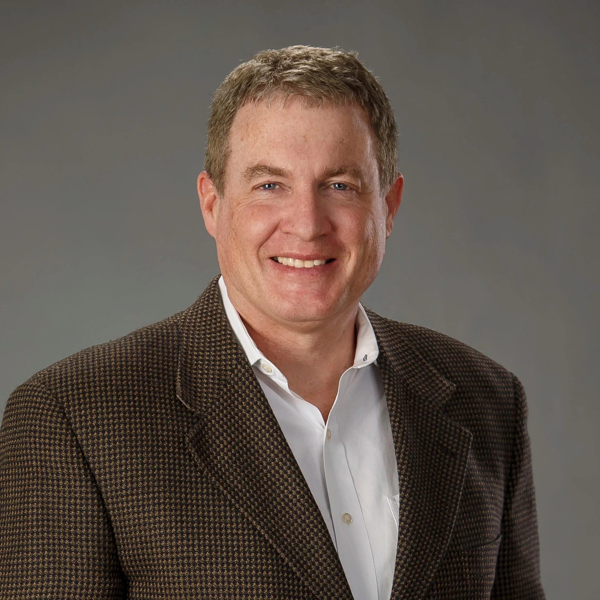 Portrait of a smiling man with short brown hair, wearing a white collared shirt and a brown patterned blazer against a gray background.