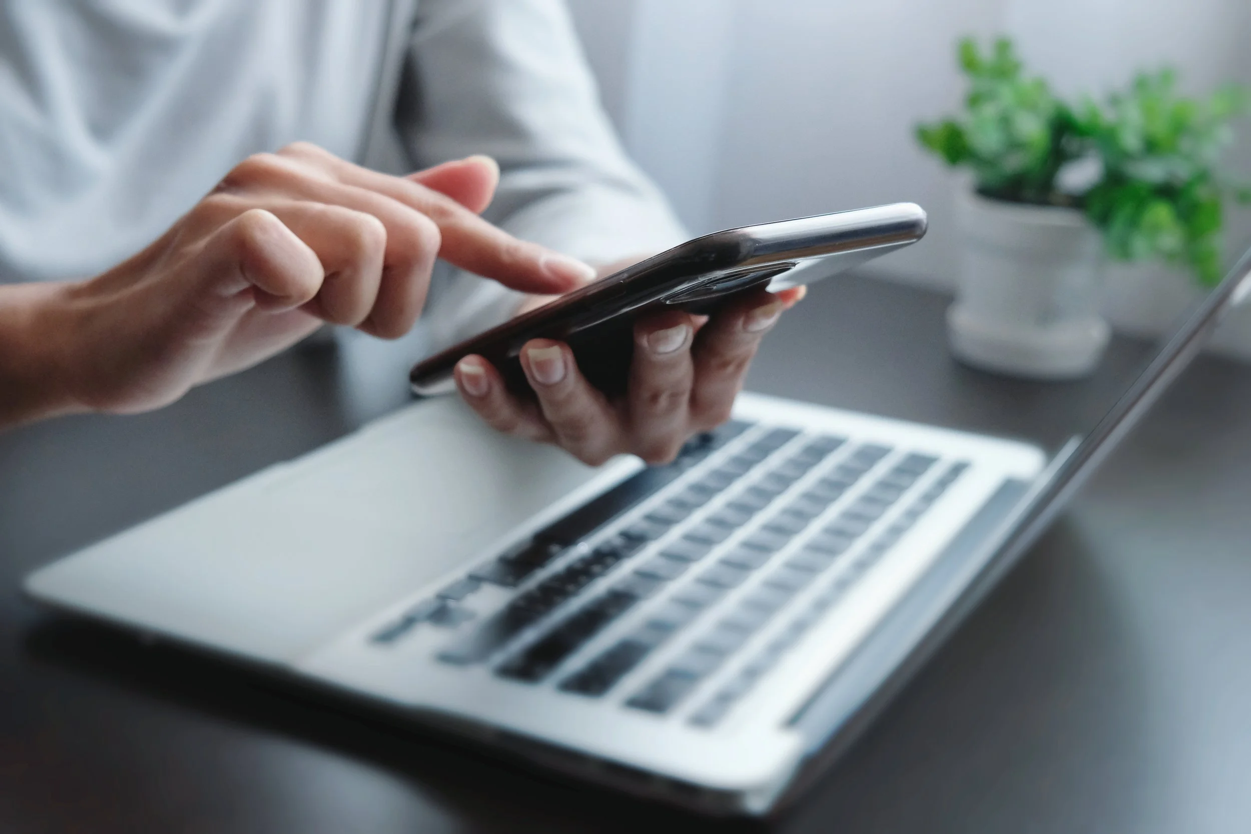 Person using a smartphone with a laptop and potted plant on a desk.