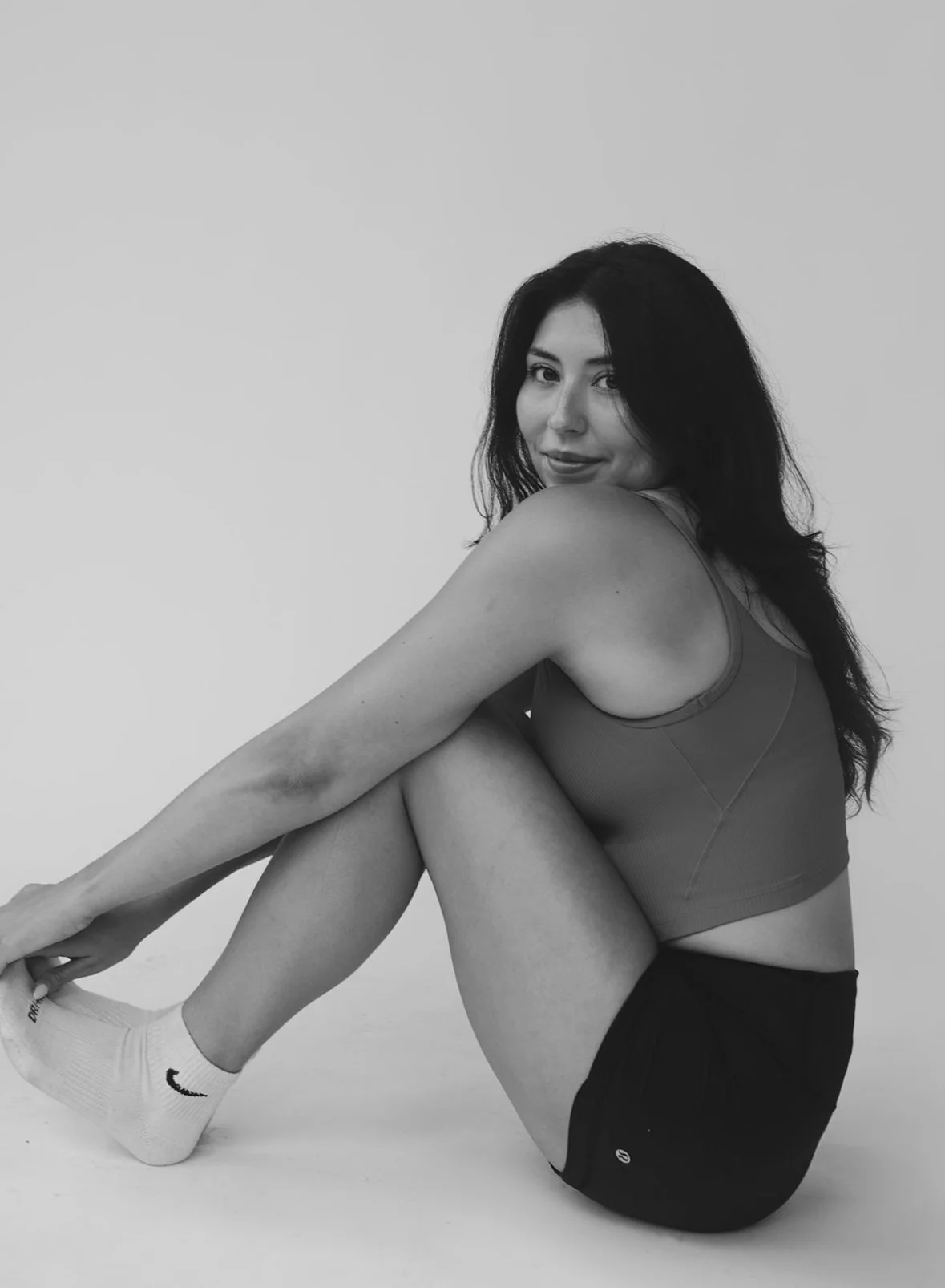 Black and white photo of a woman sitting on the floor with her legs bent, holding her foot, and smiling at the camera.