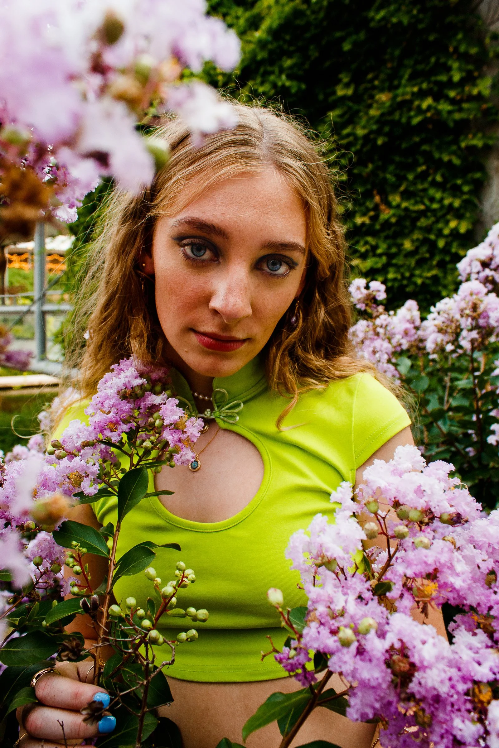 A young woman with curly blonde hair and blue eyes outdoors among pink flowers, wearing a neon green top and jewelry. Editorial model headshots, editorial, and commercial photography in St. Augustine, Florida. 