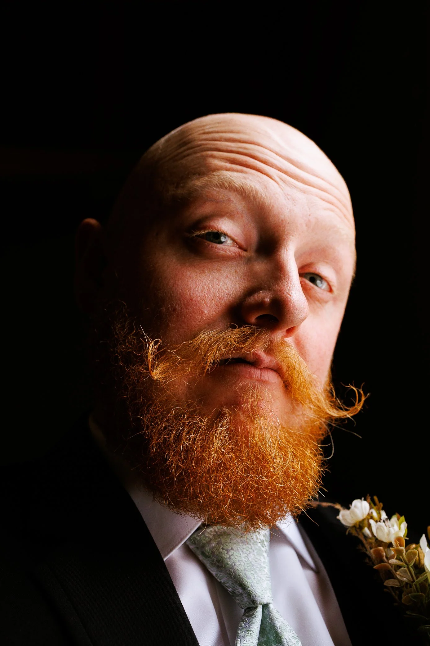 Portrait of a groom with a large red beard, bald head, and blue eyes wearing a black jacket, white dress shirt, light green patterned tie, and a boutonnière with white flowers against a black background. Fun, charismatic. St. Augustine Wedding Photo.