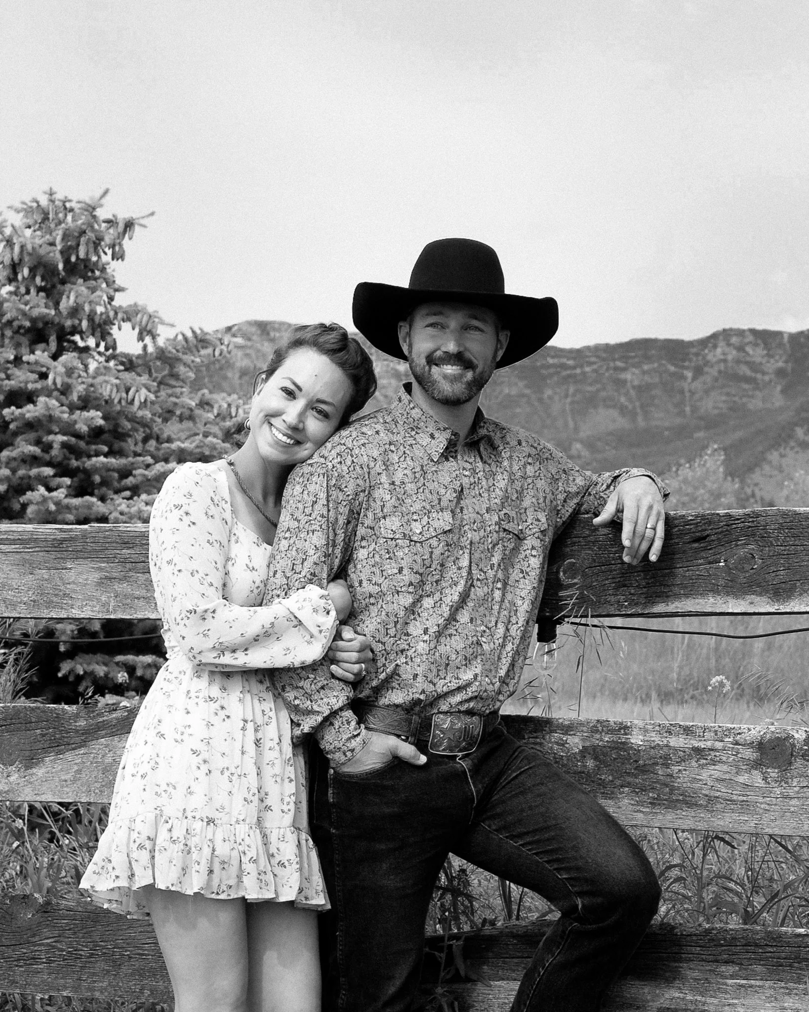 A happy couple smiling, leaning on a wooden fence outdoors, with mountains and trees in the background in a black-and-white photo. Gallatin Gateway, MT, cowboy, country, western couples portrait. By Evelyn Elizabeth Photo, Based in St. Augustine