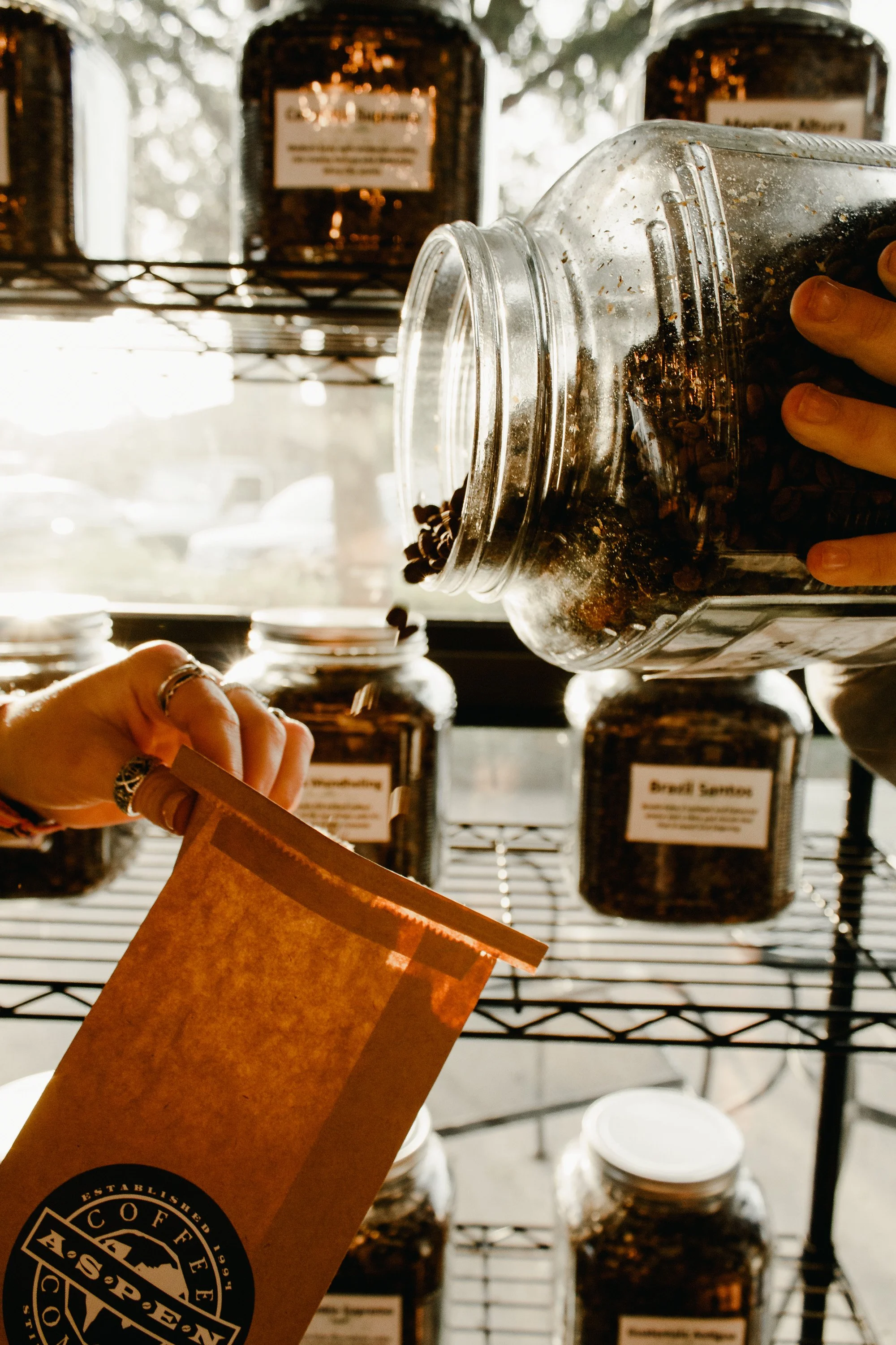 Hands holding a brown paper bag and pouring coffee beans from a glass container with other jars of coffee beans on a metal shelf behind. Warm and creative brand and small business photography in Saint Augustine, Florida by Evelyn Elizabeth Photo.