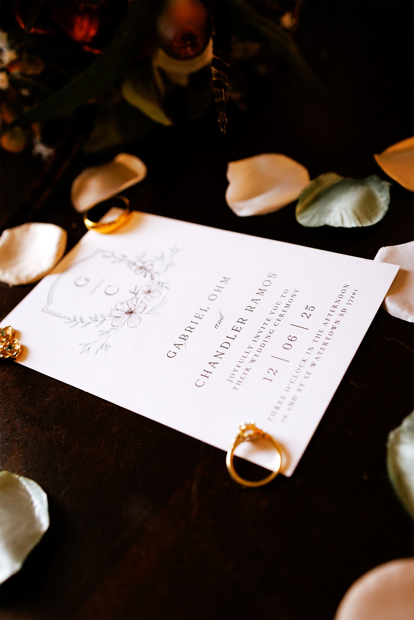 Invitation for a wedding, placed on a dark surface surrounded by gold rings and white flower petals, with greenery visible. Elegant and moody flat lay. Evelyn Elizabeth Photography, Evie Sanchez photo, St. Augustine Florida, Montana, South Dakota.