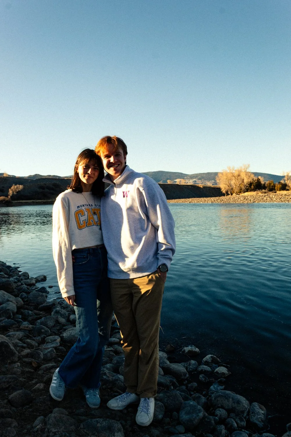 A young couple pose together by a river with a rocky shore, in a scenic outdoor setting during sunset. Golden hour, film feeling, dreamy, soft, water, cinematic couples photography in St. Augustine, Florida, Montana, South Dakota. Evelyn Elizabeth