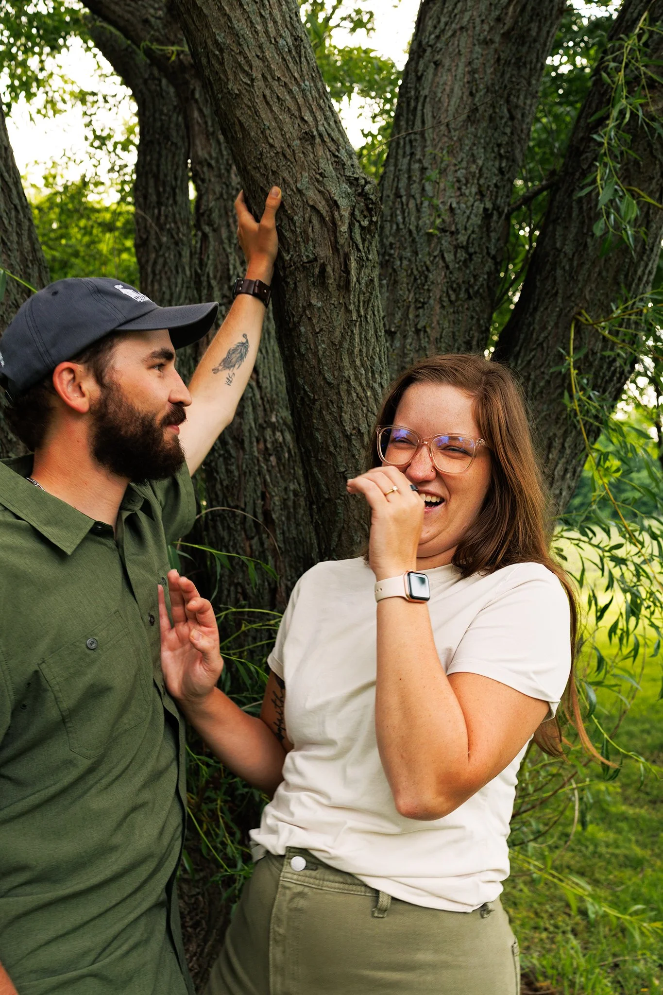 Two people stand outdoors near a large tree, laughing and engaging in a lighthearted conversation. One person, with glasses and a white shirt, appears to be amused, while the other person, with a beard and cap, is gesturing with his hand. Florida