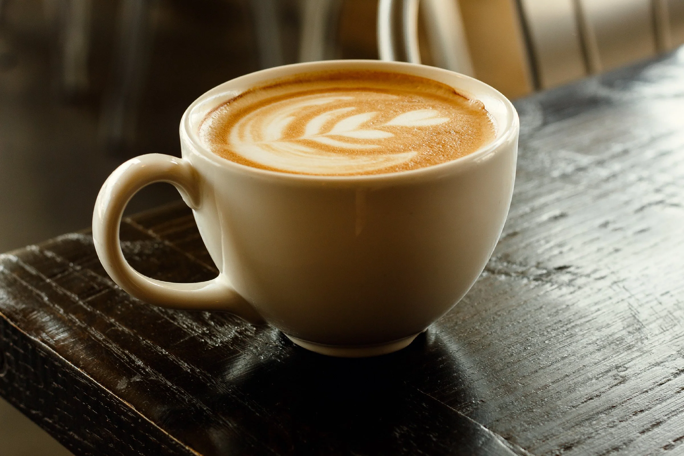A detail shot of a white ceramic mug filled with a latte displaying a leaf-shaped latte art design on top, placed on a dark wooden table. Clean and personalized restaurant content. Brand and business marketing photography in Saint Augustine.