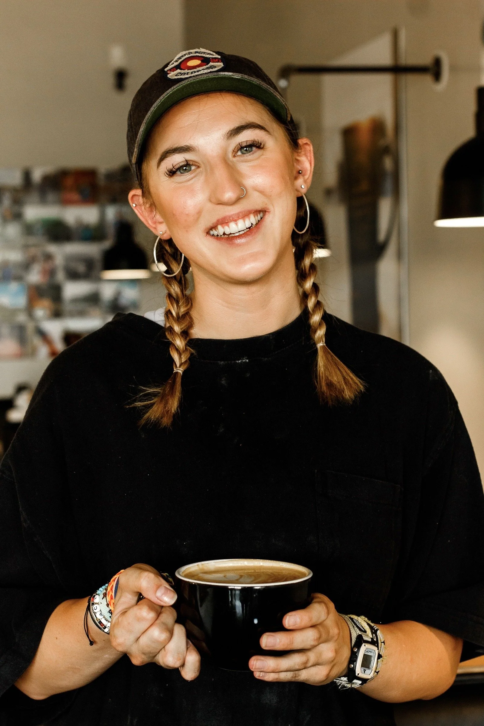 A barista/employee with braided hair, hoop earrings, and a nose ring, smiling and holding a black mug, wearing a black sweatshirt, bracelets, and a watch, in a cozy indoor setting. Sharp marketing and content photography in St. Augustine, FL