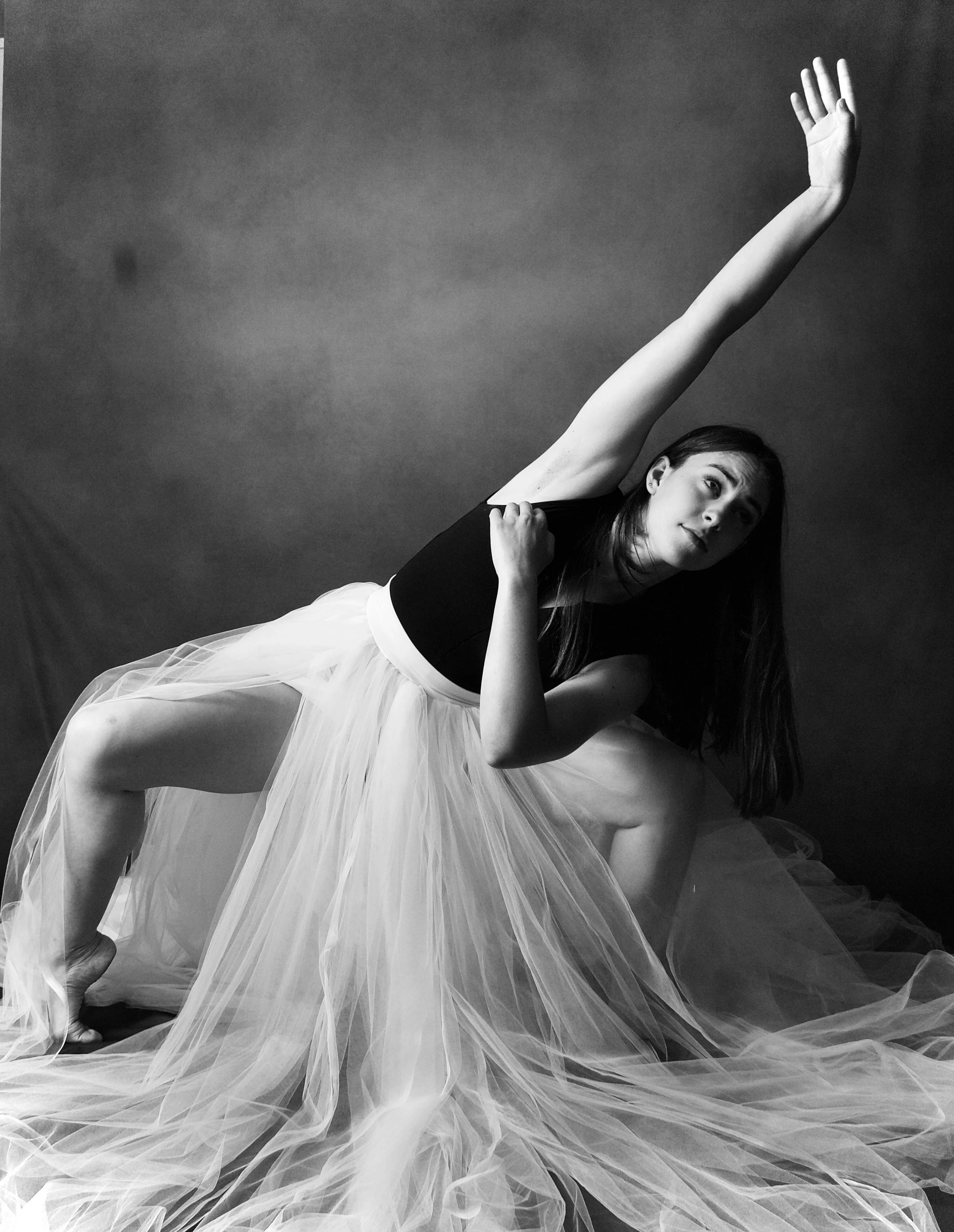 A ballerina wearing a black top and a flowing white tulle skirt is posing on a plain background, with her arm raised and a gentle expression on her face in b&w. Timeless sports, dance, editorial, commercial photography in St. Augustine, Fl.