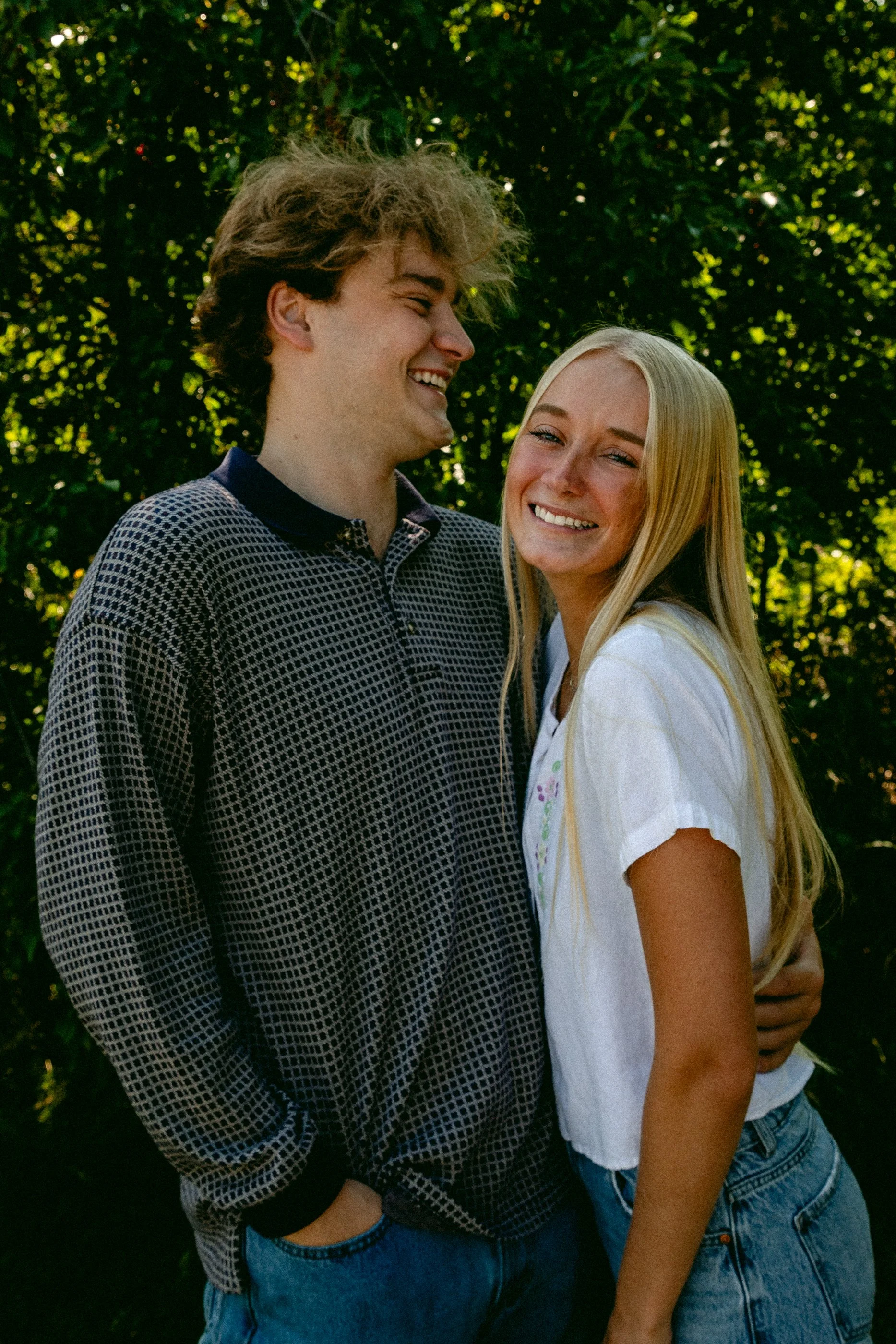 A young man with curly hair and a young woman with long blonde hair smiling and standing close together outdoors with greenery in the background. Sharp, colorful, dreamy photograph. Montana, South Dakota, Florida couples portrait.