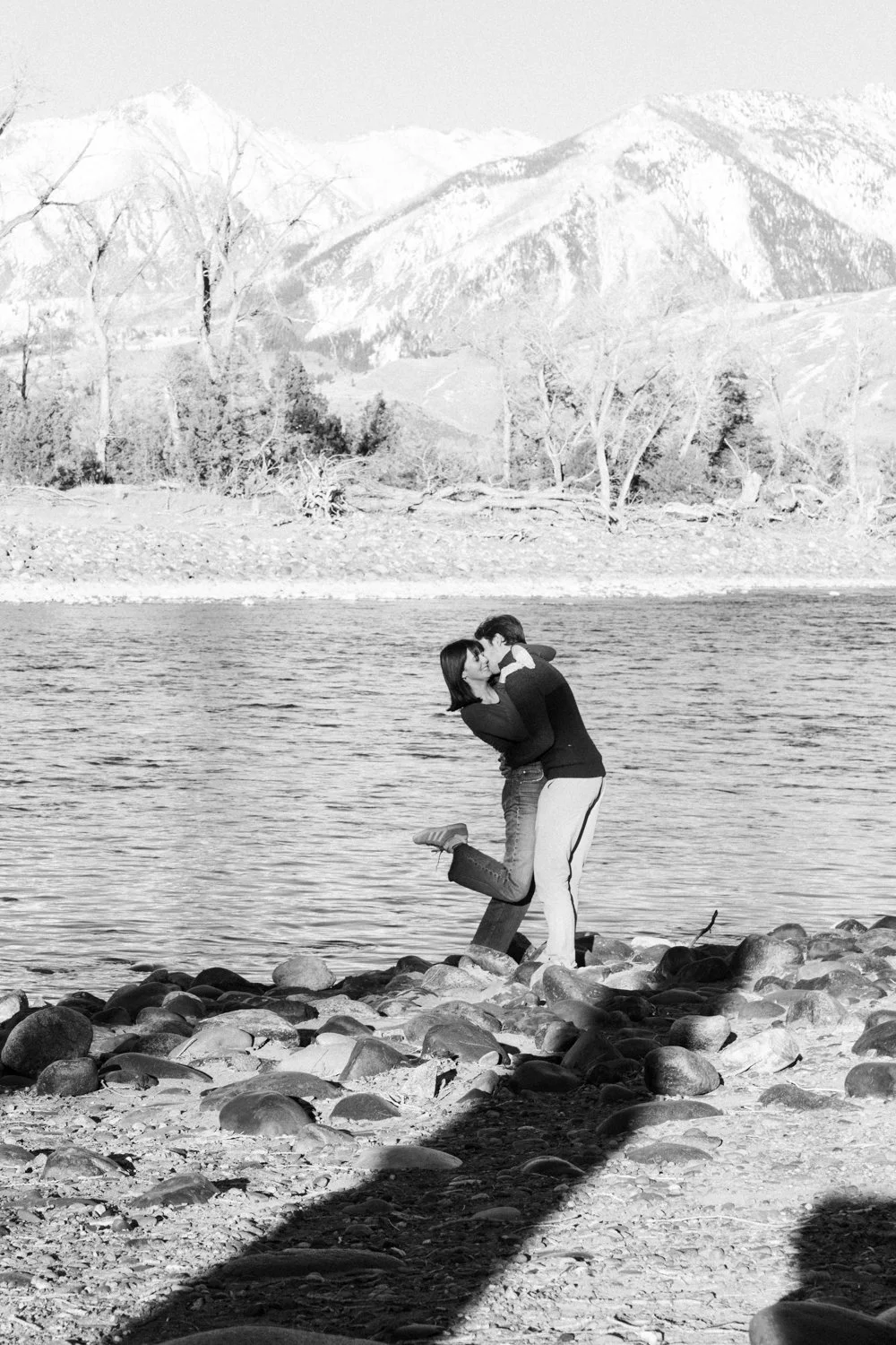 A picturesque scene of a couple  kissing on rocks by a river, with mountains and trees in the background. Black and White, scenic, outdoors. Dreamy, memory, sweet couples and engagement photography in St. Augustine, Florida. Affordable. By Evelyn