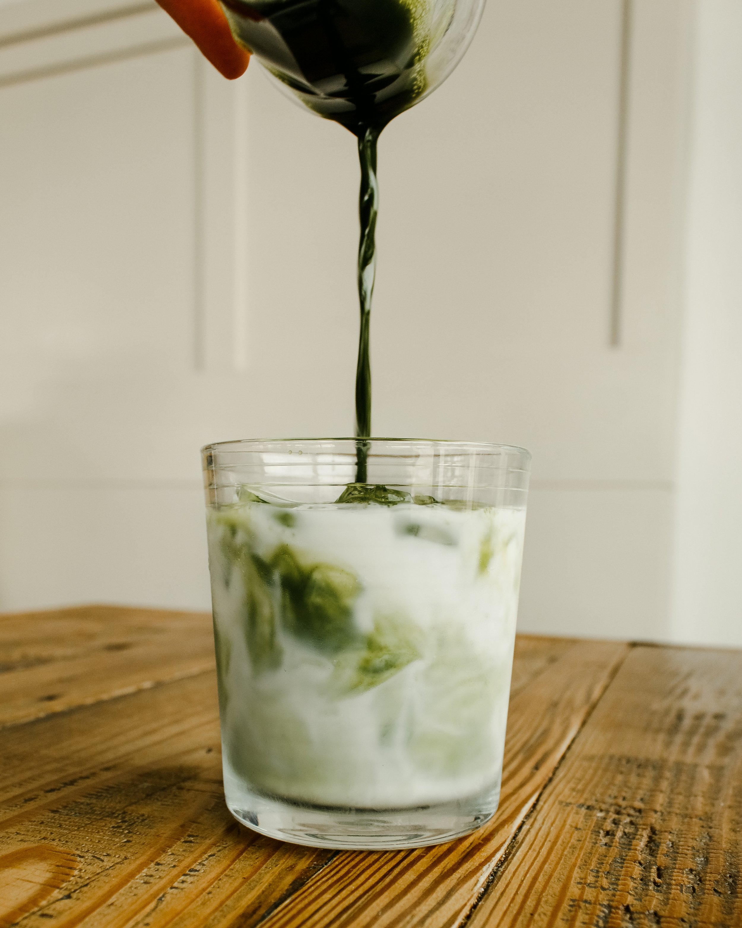 A hand is pouring green iced matcha latte from a glass pitcher into a tall glass filled with ice on a wooden table. The background is simple and neutral. Clean brand and business marketing photography by Evelyn Elizabeth photography based in St. Aug.