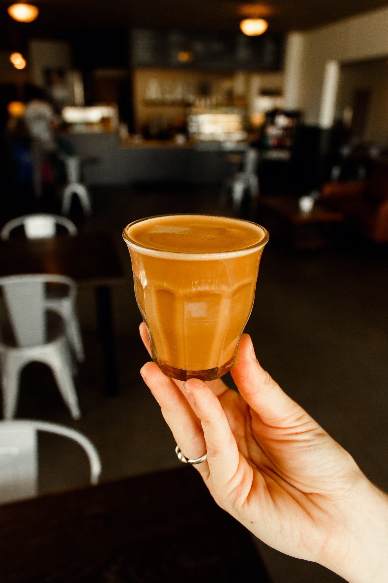 A hand holding an espresso shot glass  in a cafe setting with blurred background of chairs and tables. Professional brand content, marketing, and event photography in St. Augustine, Florida by Evelyn Elizabeth Photography.