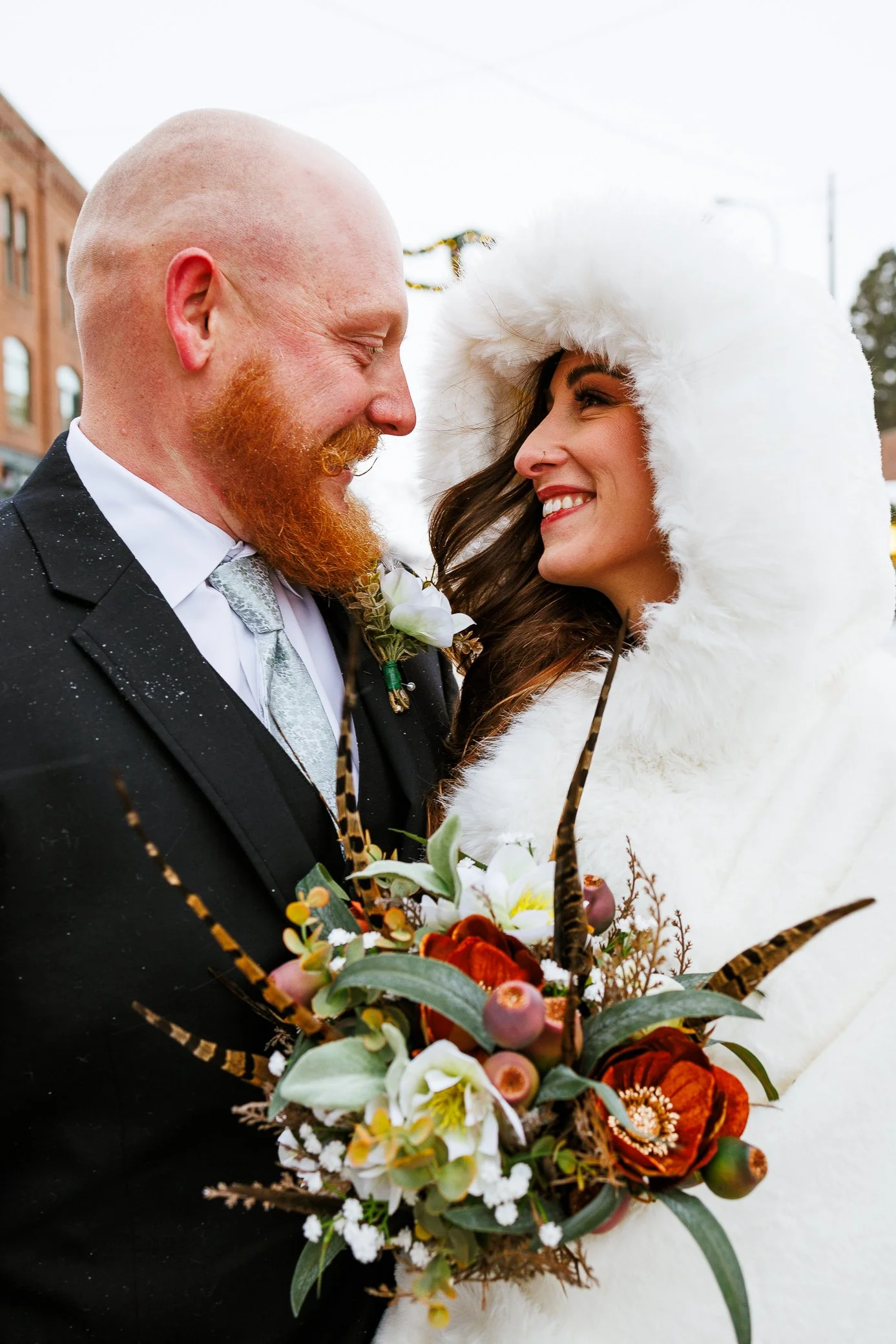 Candid portrait of a bride and groom smiling at each other, the bride wearing a white fur-lined coat, and holding a bouquet of flowers with feathers and greenery. Dreamy, intimate, popular, trendy, affordable wedding photography in St. Augustine, Fl.
