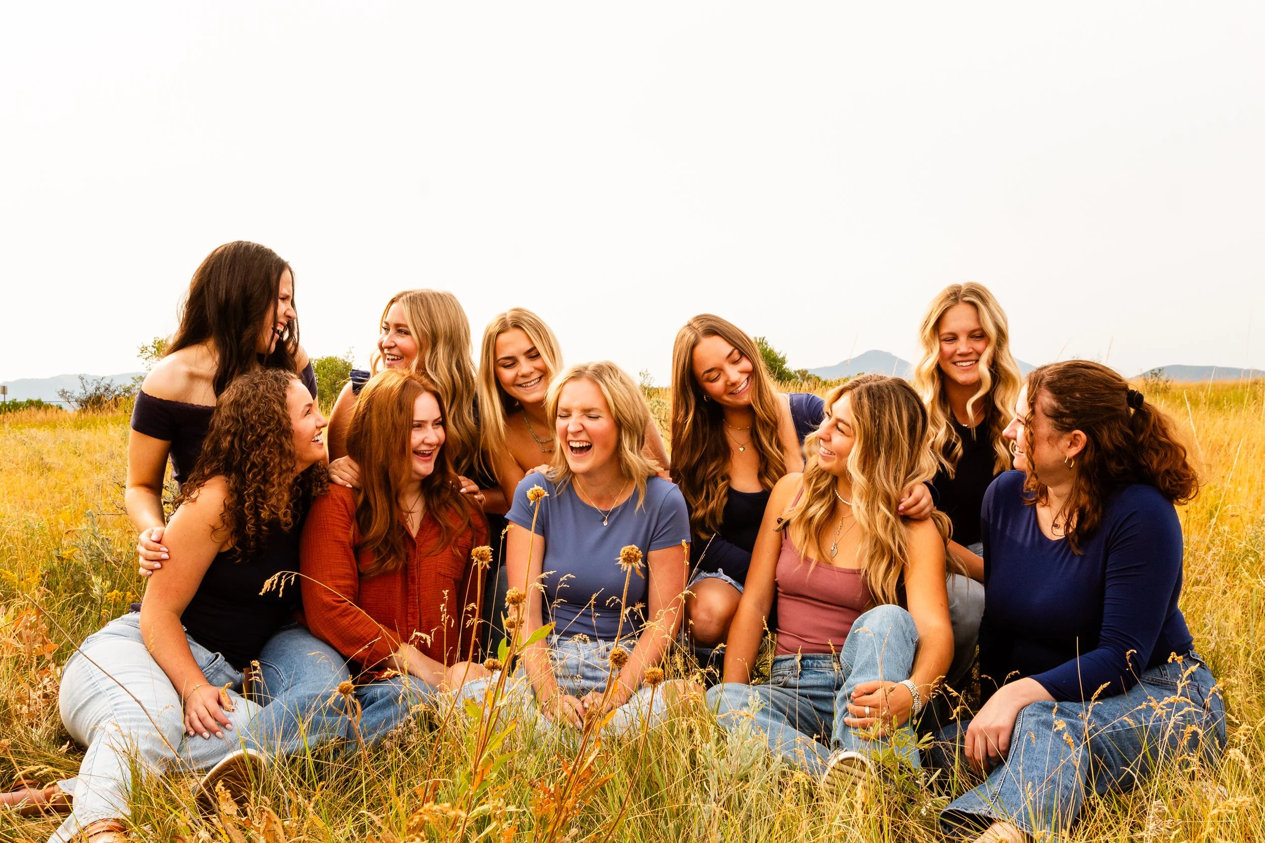 Candid group of smiling women sitting and laughing in a grassy field with mountains in the background. Professional and warm photography of organizations and teams. Genuine and personal brand photography in St. Augustine, Fl.