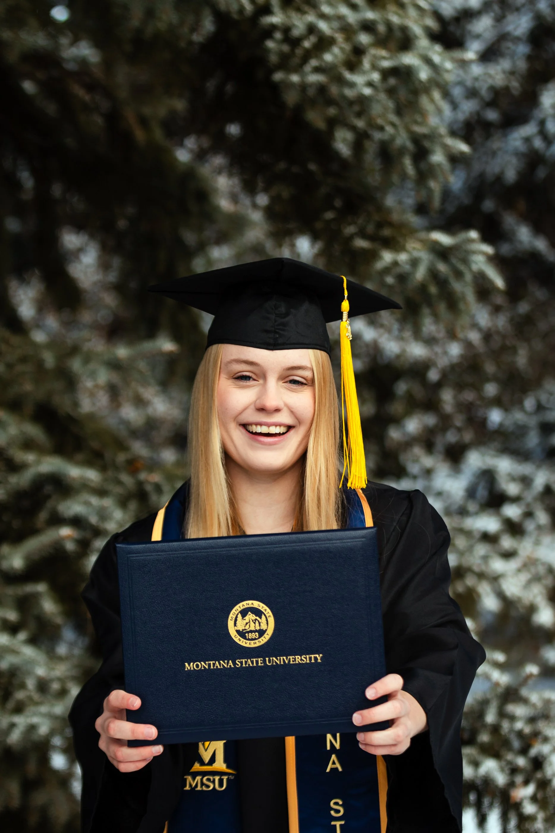 A college senior smiling, wearing a black graduation cap and gown with yellow accents, holding a Montana State University diploma cover in front of her, standing outdoors with snow-covered trees in the background. Florida, South Dakota Photography.