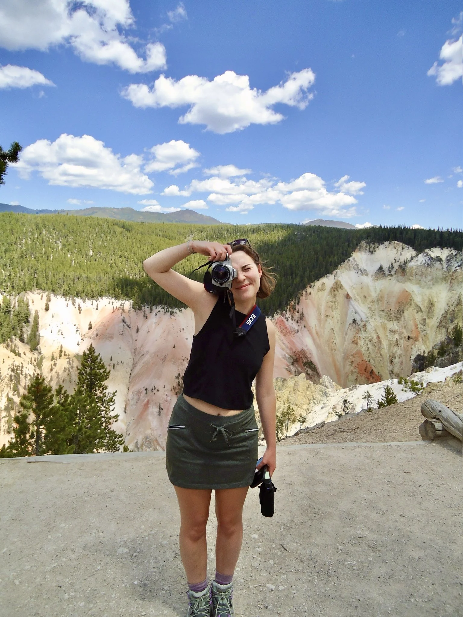 Evie Sanchez, aka Evelyn Elizabeth Photography, taking a 35mm film photograph on a dirt path in front of the Lower Falls of Yellowstone with trees, mountains, blue sky, and white clouds in the background. Evelyn is a professional working photographer