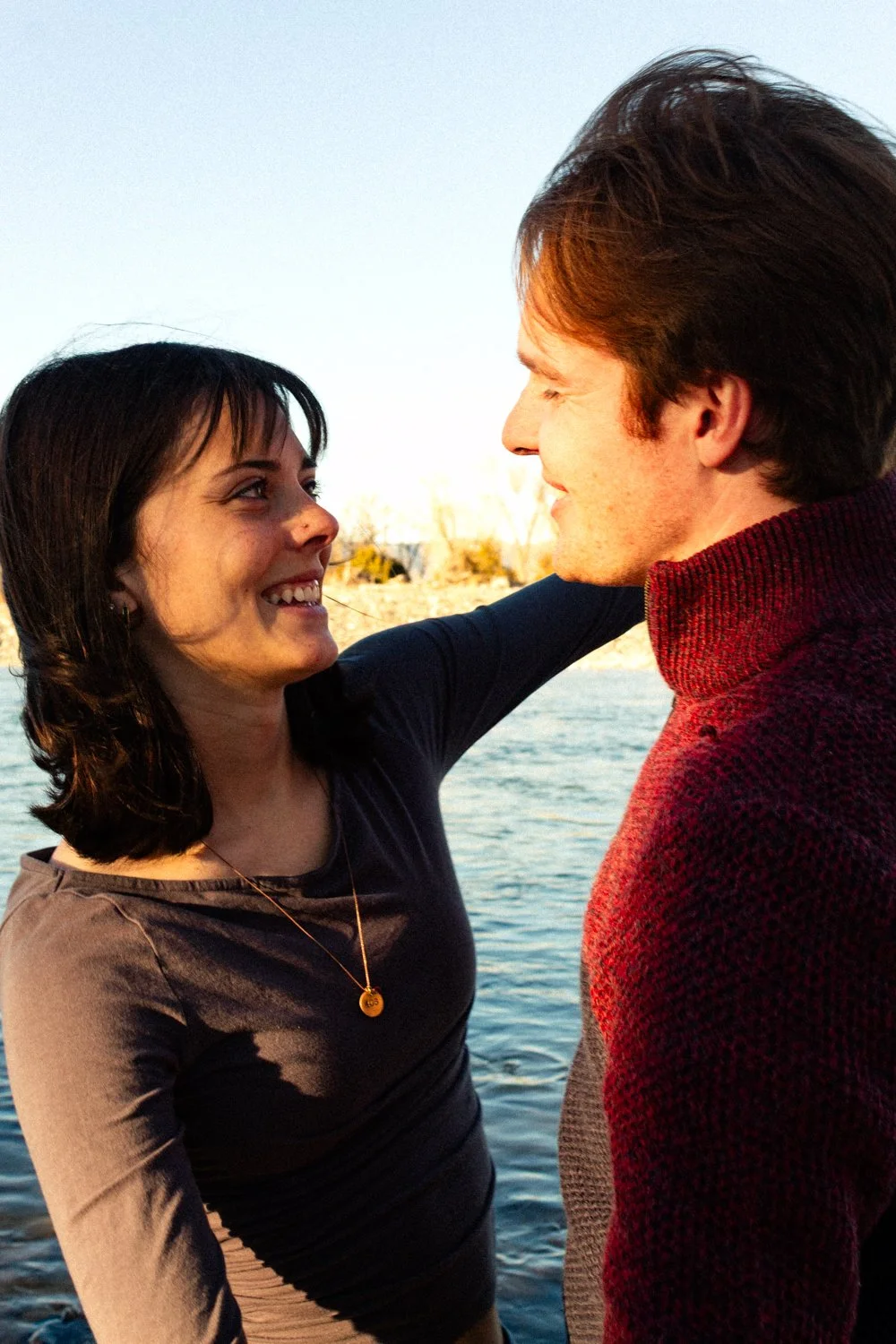 A woman and a man face each other closely by a body of water in Paradise Valley in Montana, smiling at each other romantically. Warm dreamy tone, soft texture, intimate feeling, genuine mood. Couples photography in Montana, Florida, South Dakota.