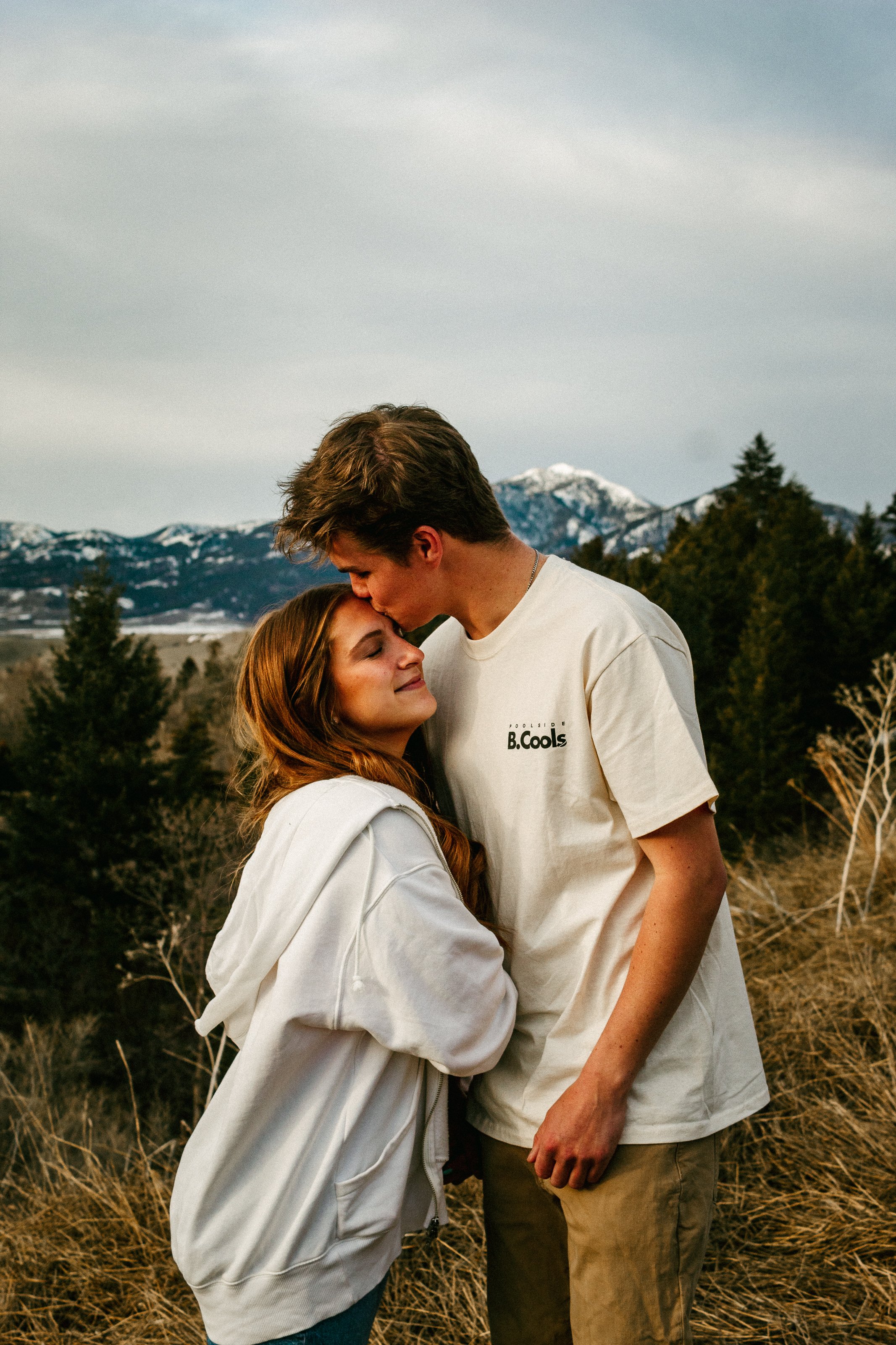 A young couple standing outdoors in a mountainous area with snow-capped peaks and pine trees, sharing a tender moment with the boyfriend kissing the woman's forehead as she closes her eyes. Dreamy, warm, outdoors, mountain, romantic, genuine, simple.