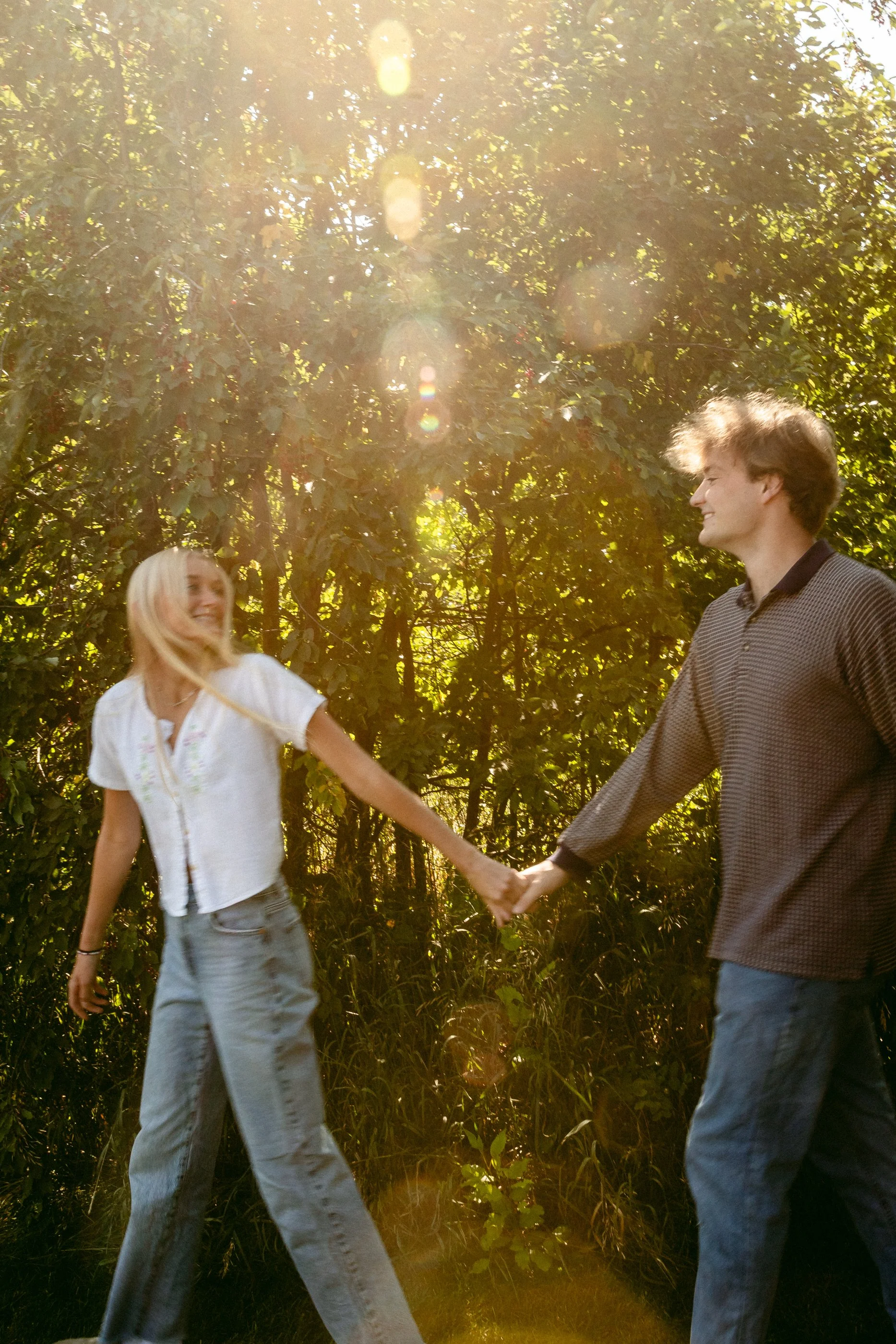 A young man and woman holding hands outdoors in a wooded area during sunlight. A motion photograph with golden sun spots and green foliage. Romantic, candid photography with genuine laughter. Montana, South Dakota, Florida  couples portrait.