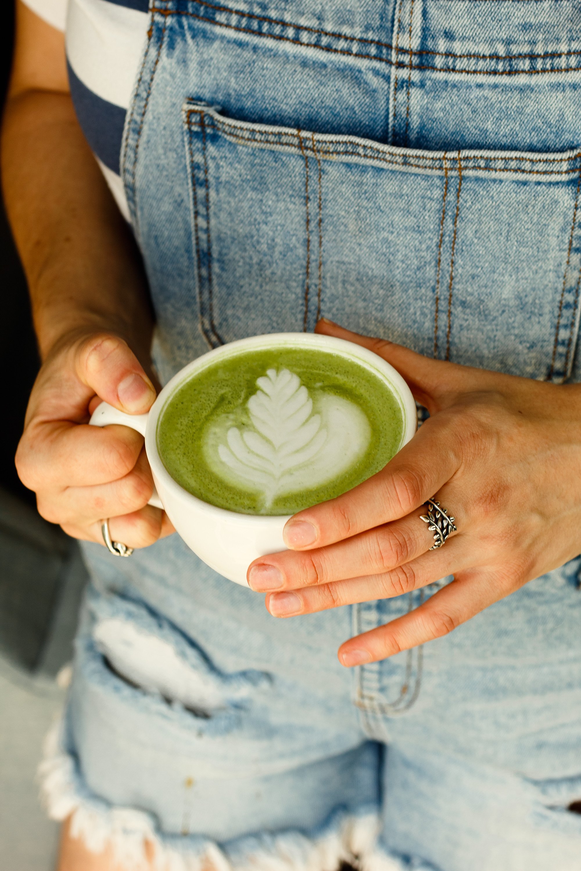 Social media content: a customer holding a white ceramic mug of a green matcha latte with latte art in the shape of a flower, wearing a denim apron over a striped shirt. Clean brand photography in St. Augustine, FL by Evelyn Elizabeth Photography.
