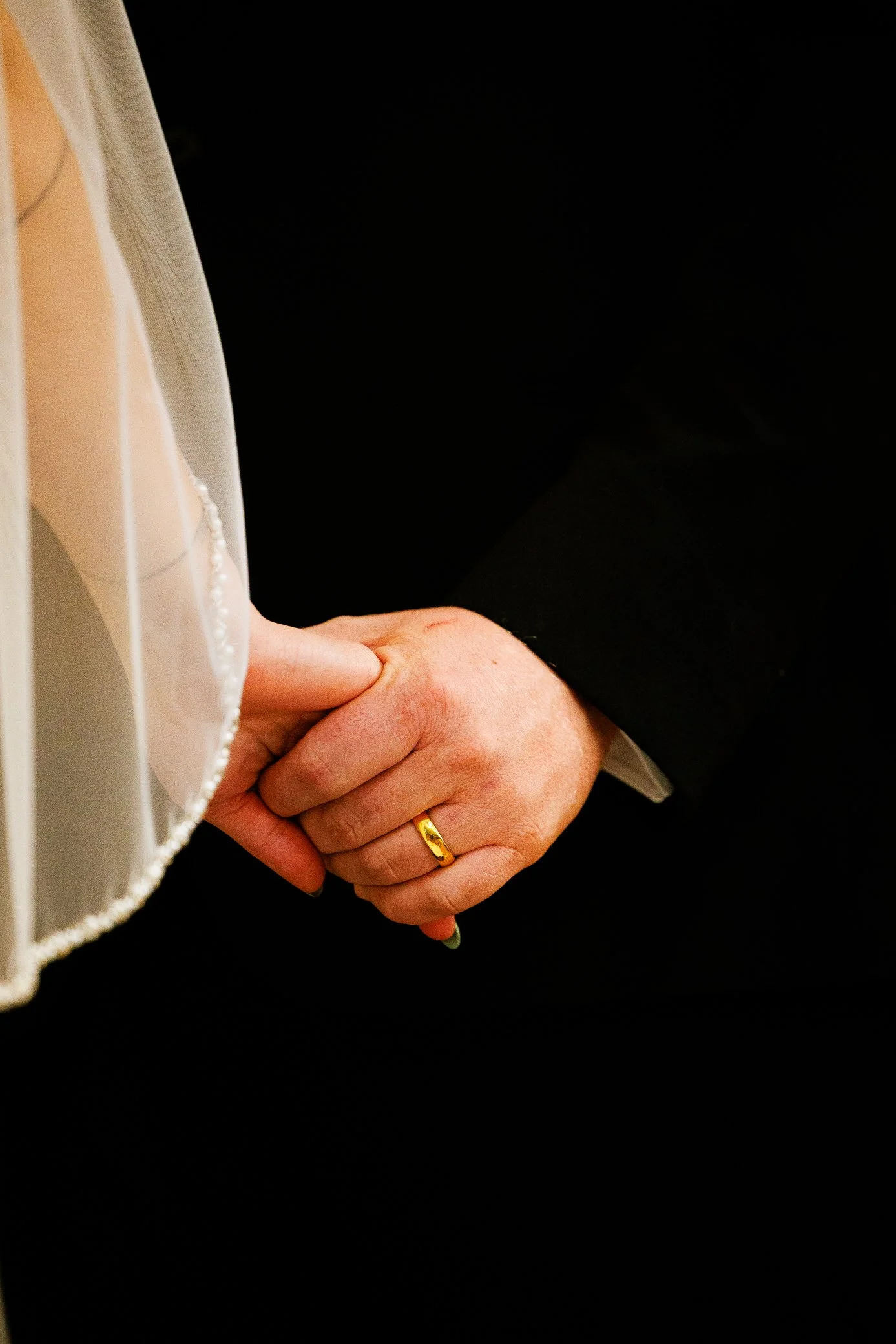 A close-up detail of a bride and groom holding hands during their wedding ceremony, with the bride wearing a veil and a white dress, and the groom dressed in a black suit. Dreamy, light, moody, brightMontana, South Dakota, Florida  wedding photo.