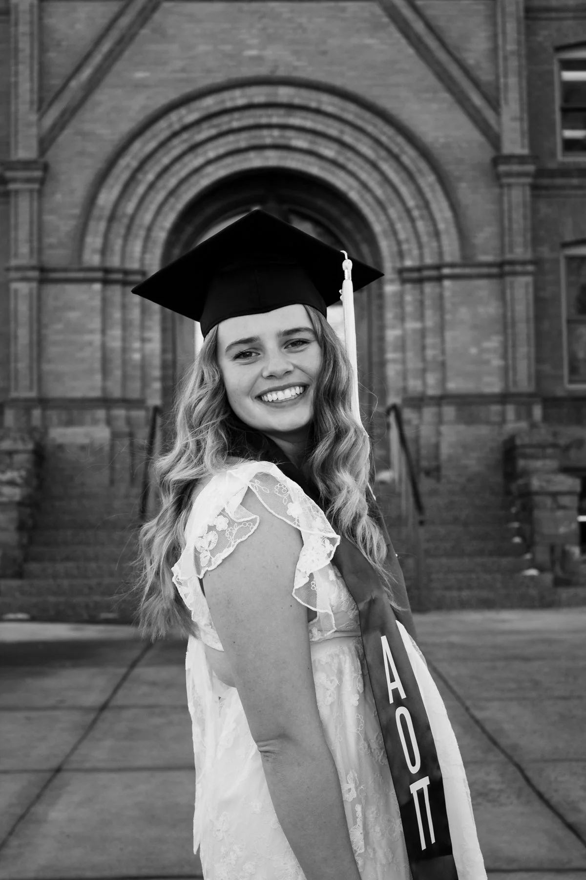 A young woman in a graduation cap and gown smiling in front of a historic brick building with arched windows and steps. Montana State University, Flagler College, and UF senior graduation popular photographer. Trendy and timeless in St. Augustine.