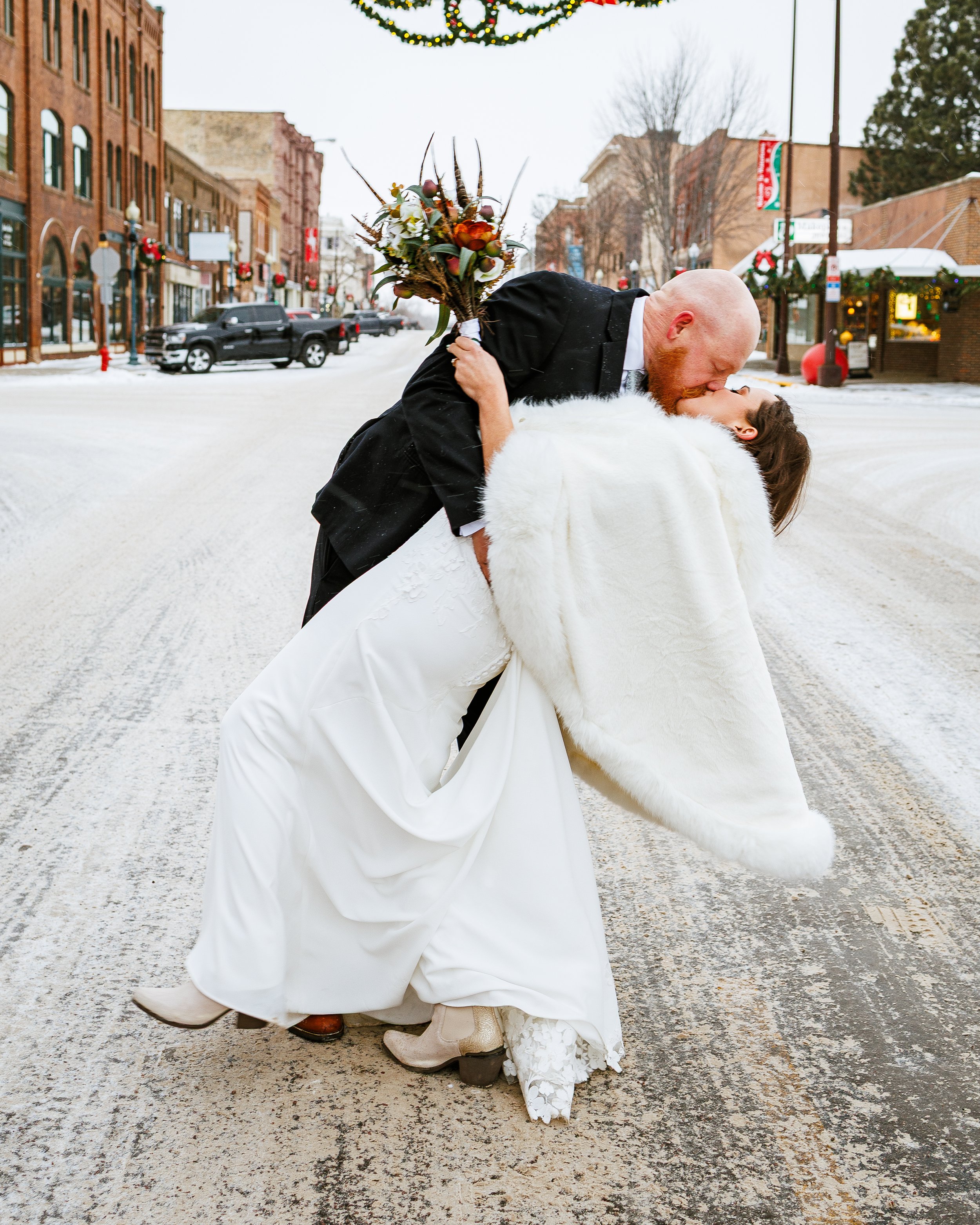 Newlyweds dressed in formal wedding attire romantically kissing on a snowy street decorated with Christmas wreaths and lights. The bride is dipping backwards in the groom's arms, holding a bouquet of flowers. Festive, dreamy, elegant, timeless.