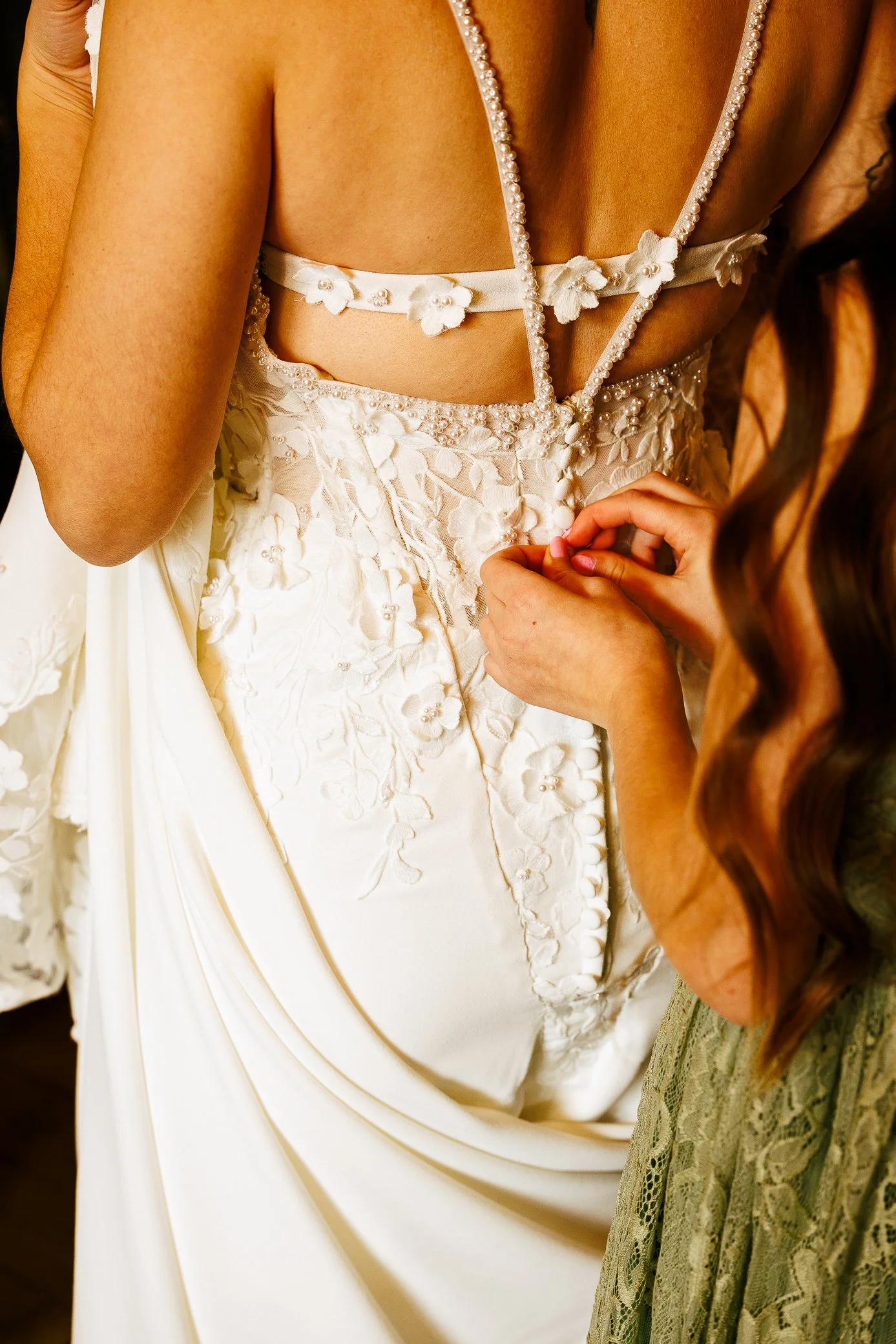 Dreamy candid detail photography of a bride getting ready with the help of her bridesmaid. Elegant, timeless structure with pearls and flower embellishments on flowy white wedding dress. Wedding and engagement photography in St. Augustine, Florida.