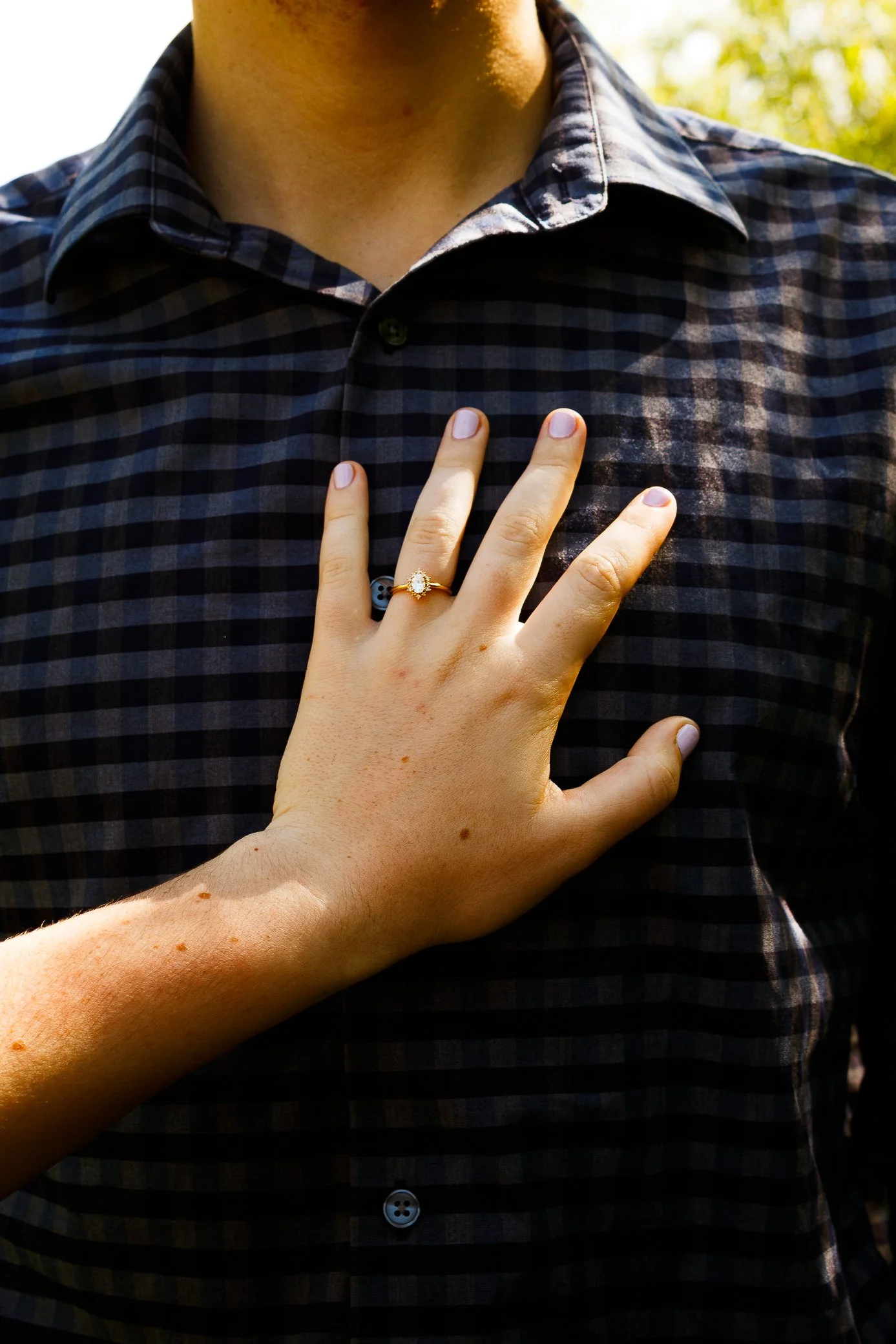 A fiancé wearing a dark plaid shirt, with a hand showing an engagement ring placed on the chest. Engagement photography. Warm, soft texture/grain, dreamy, romantic, green, summer. High quality couples photography in Florida, Montana, South Dakota.