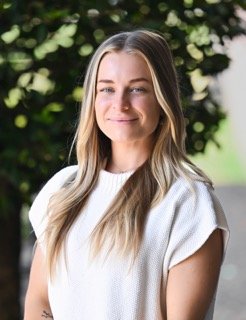 A young woman with long blond hair, wearing a white top, smiling softly outdoors with greenery in the background.
