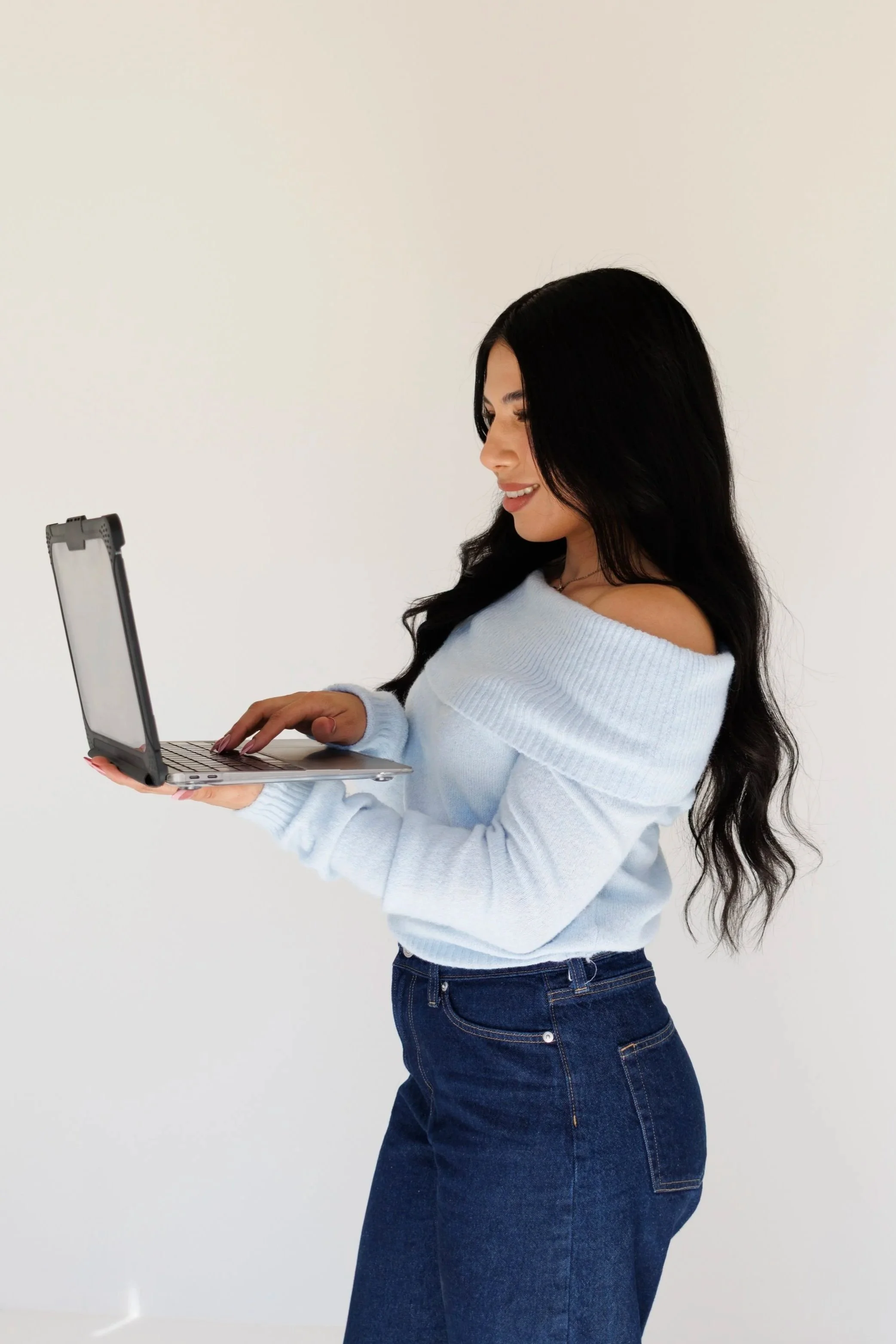 A woman with long dark hair smiling while looking at her laptop against a plain white background.