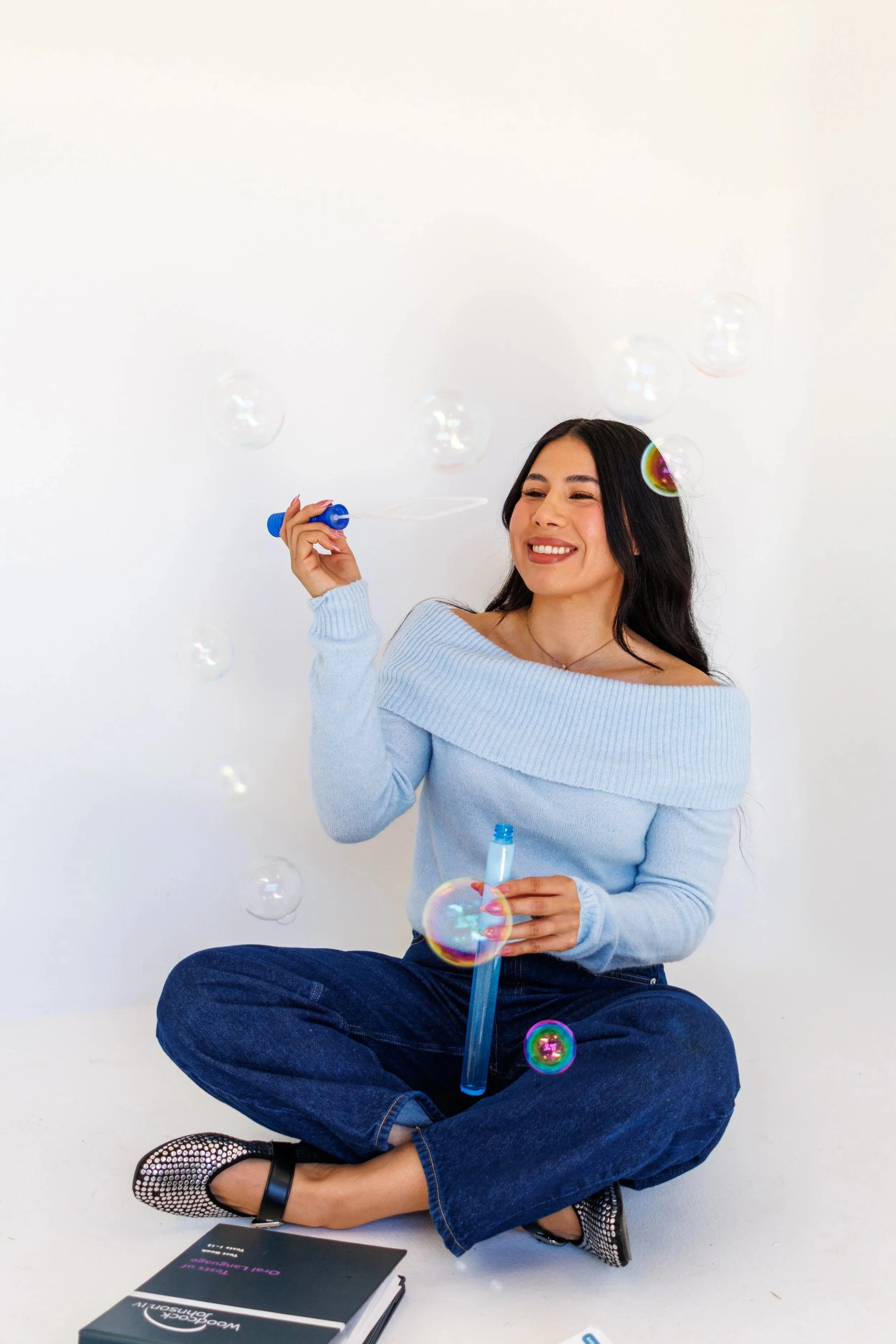 A woman sitting cross-legged on the floor, smiling and blowing bubbles with a bubble wand. She is wearing a light blue off-the-shoulder sweater, dark blue jeans, and patterned shoes. There is a black notebook or folder with a pen on the floor beside her, and a plain white background.