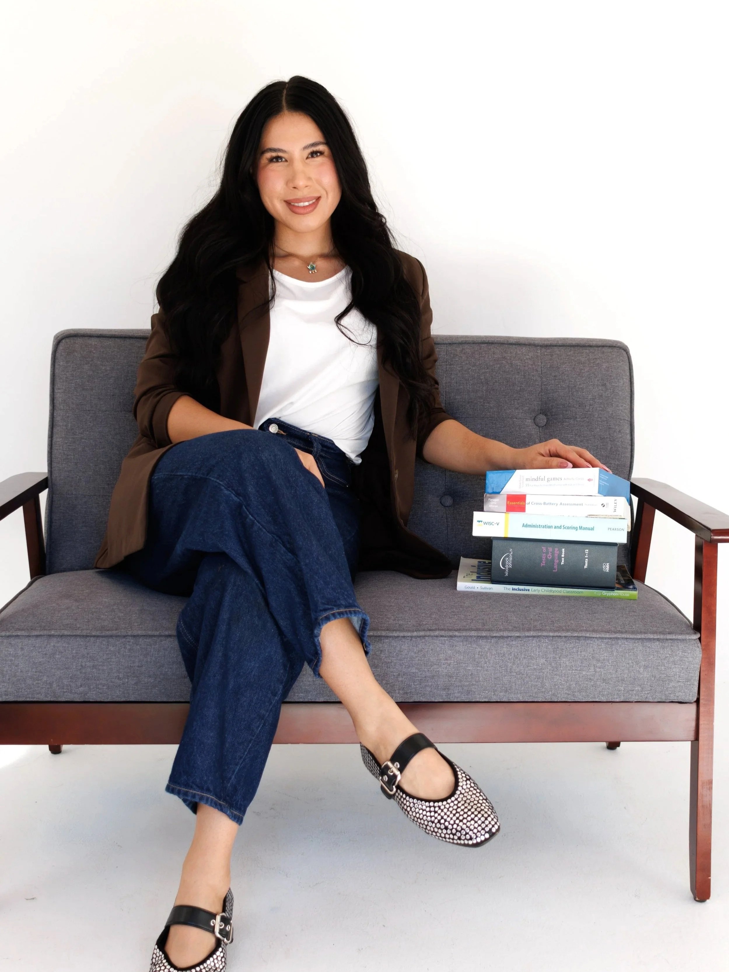A woman with long black hair, sitting on a gray sofa, smiling, wearing a white t-shirt, brown blazer, blue jeans, and black-and-white shoes, with a stack of books beside her.