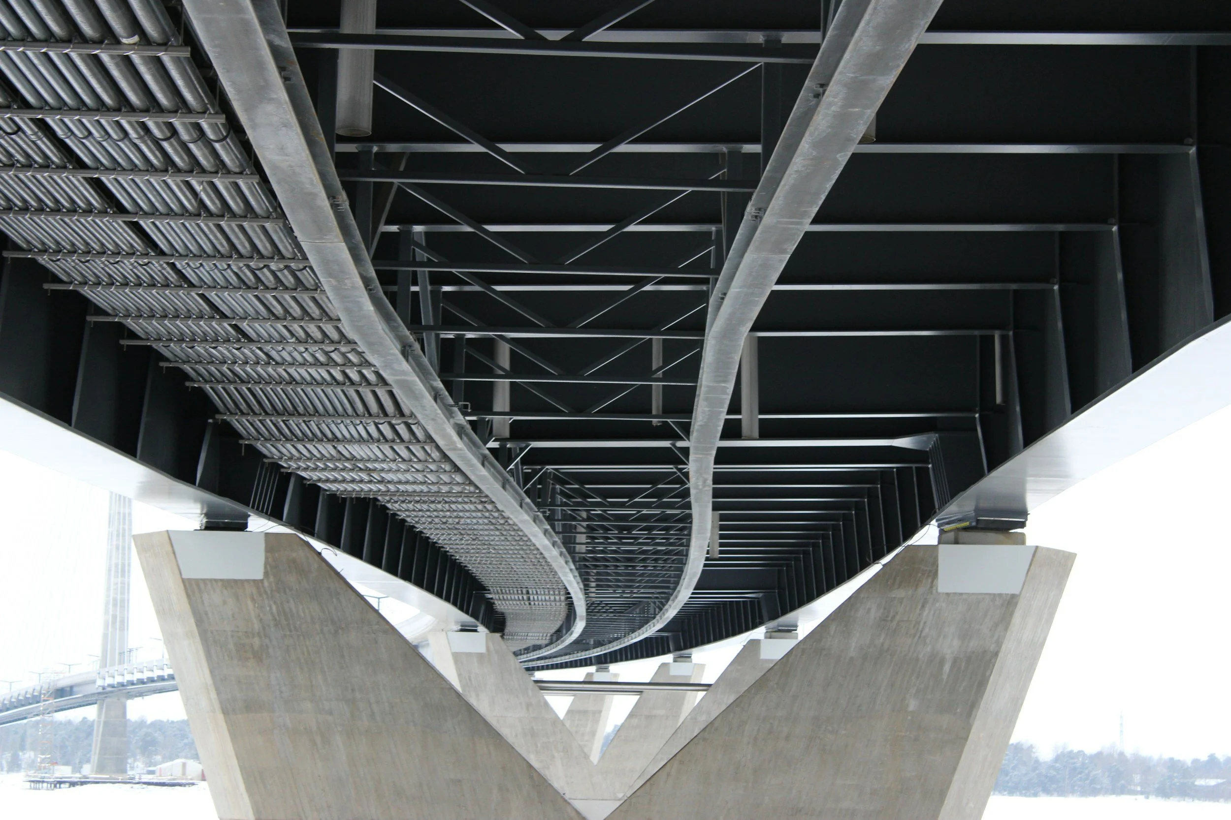 Underneath view of a modern suspension bridge showing black and gray support cables, metal beams, and concrete pillars with a snowy landscape in the background.
