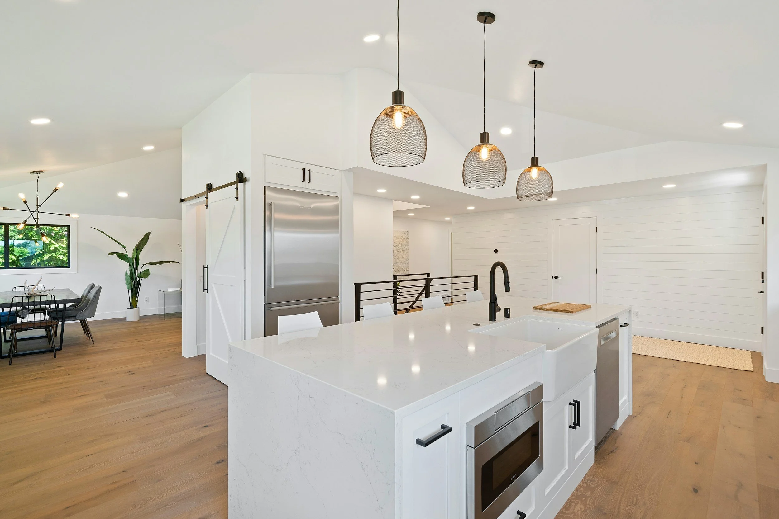 Modern kitchen with white cabinetry, a large island with a white marble countertop, pendant lights, black hardware, stainless steel appliances, and an open dining area with wooden floors and large windows.