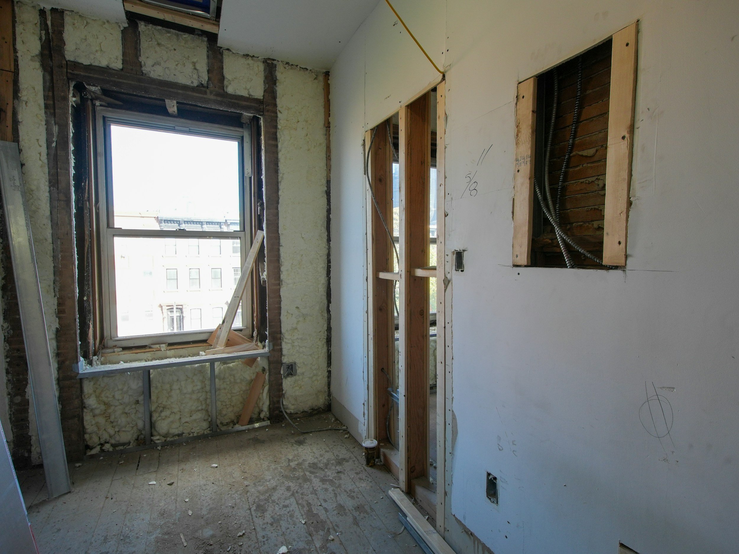 Interior of a room under construction or renovation with exposed studs, electrical wiring, insulation, and unfinished walls, with a window showing a building in the background.