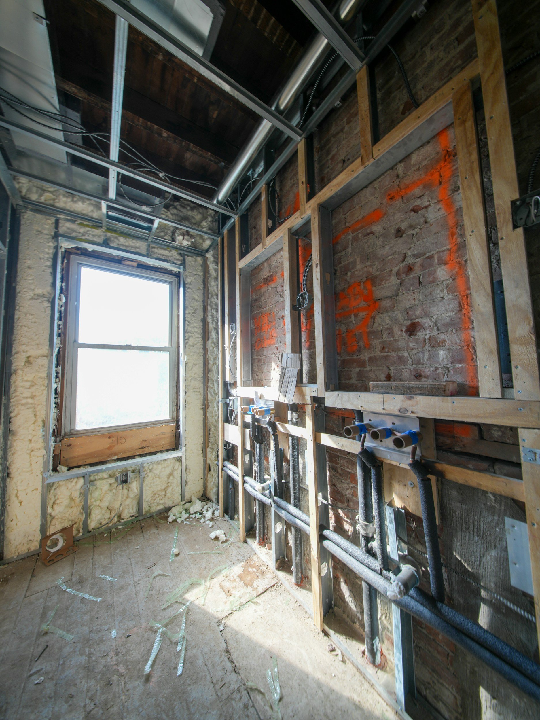 Interior of a room under construction with exposed brick and wooden framing, plumbing pipes, and a window.