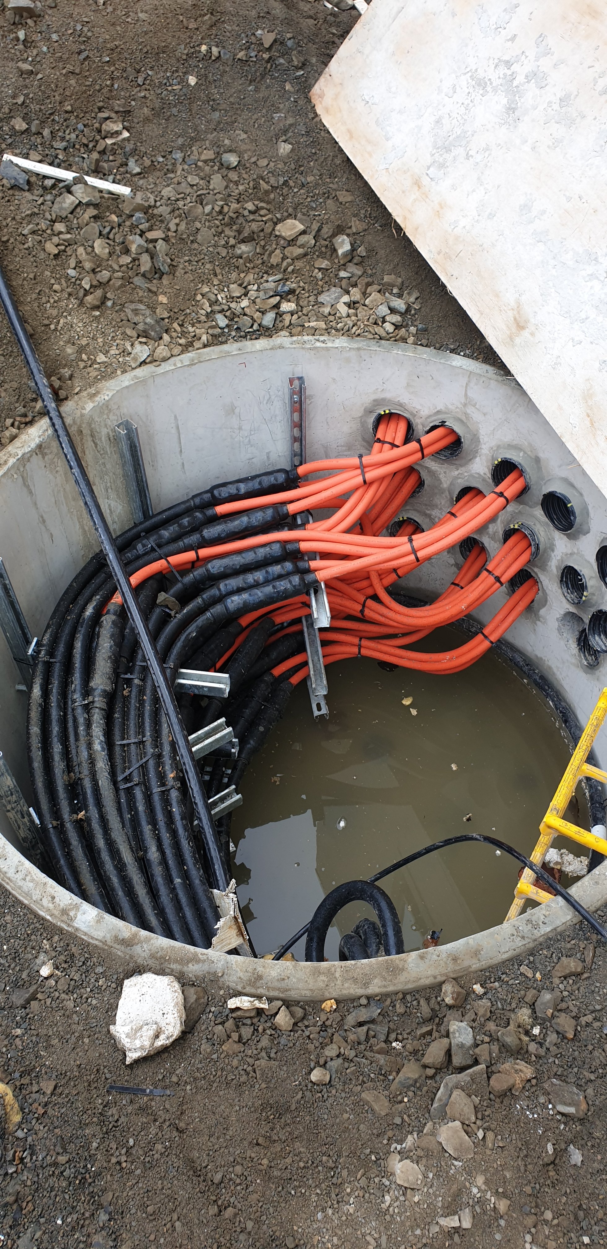 Underground utility access with orange and black cables, some in conduits, inside a circular concrete structure, with water pooled at the bottom, and gravel and dirt surrounding it.