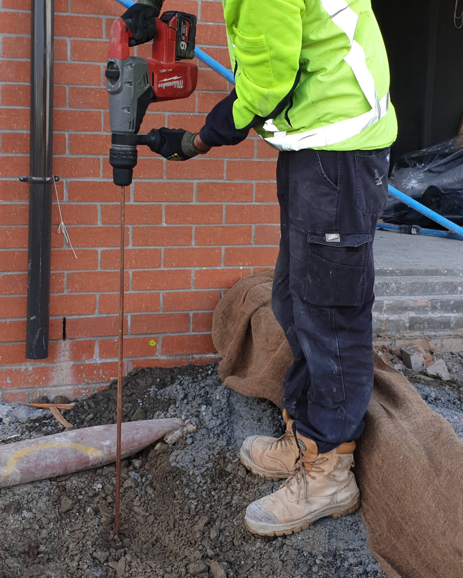 Worker in a high-visibility jacket and safety boots using a power drill at a construction site next to a brick wall.