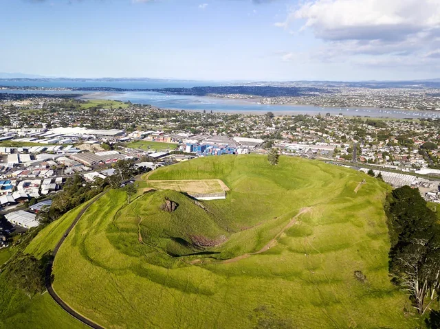 Aerial view of a green, grassy hill with an ancient giant carved into the hillside, overlooking an urban area and a large body of water in the background.