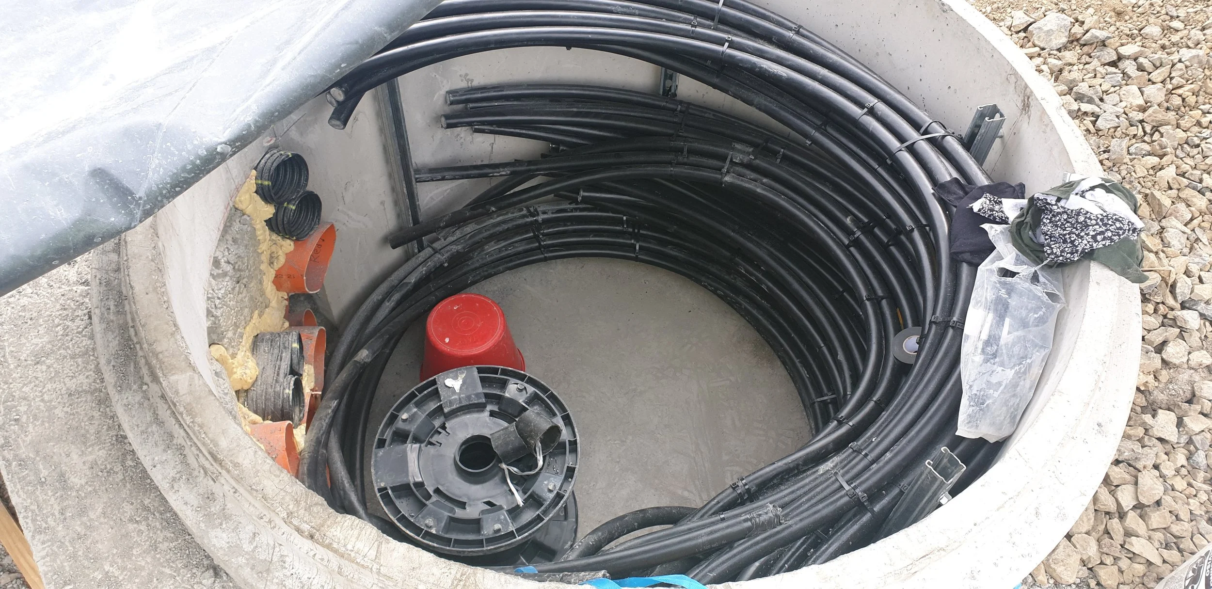 An open underground utility manhole containing black cable pipes, with some construction materials and debris around the opening.