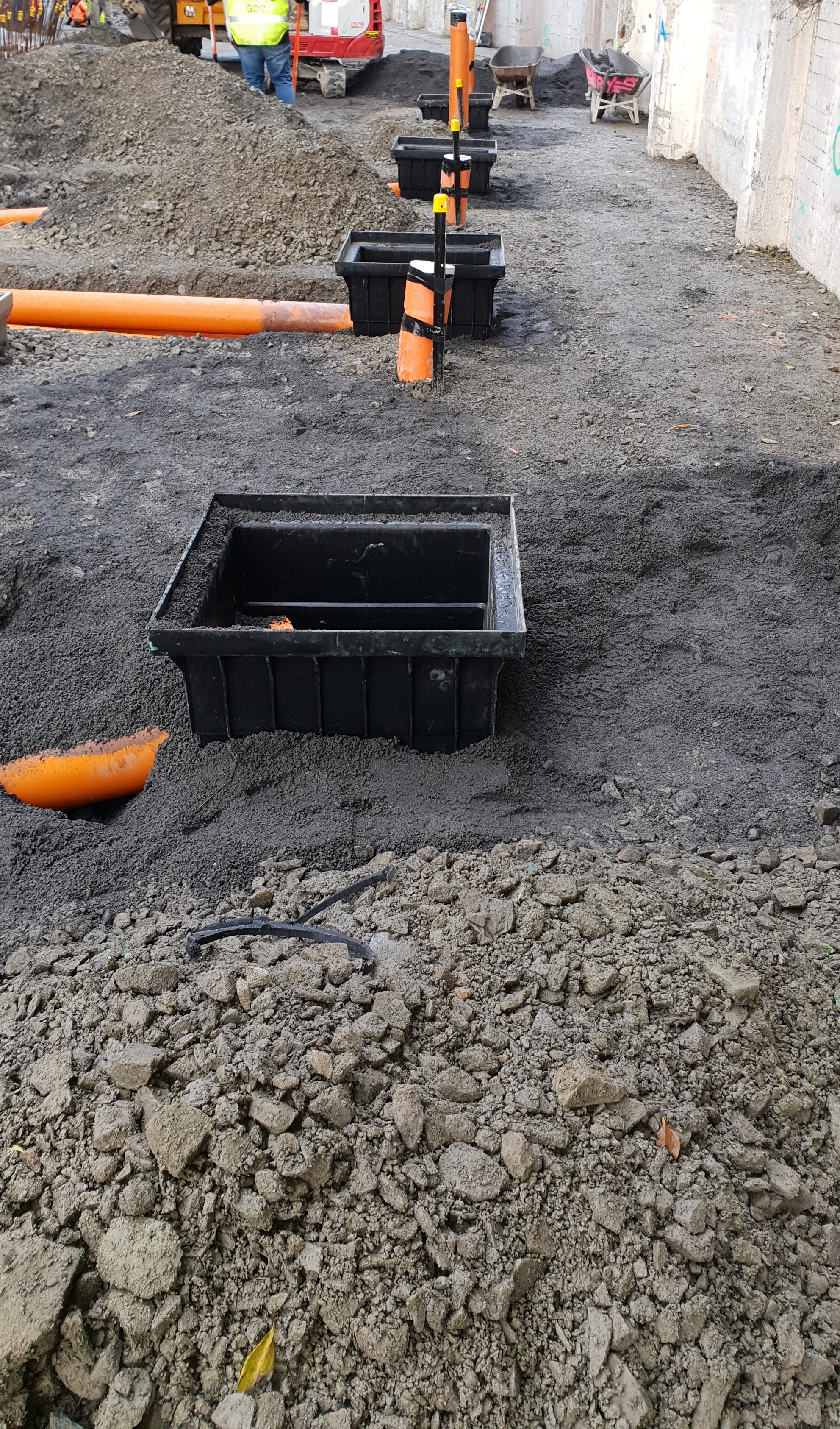 Construction site with open trenches, black plastic utility boxes, orange pipes, and construction workers in the background.