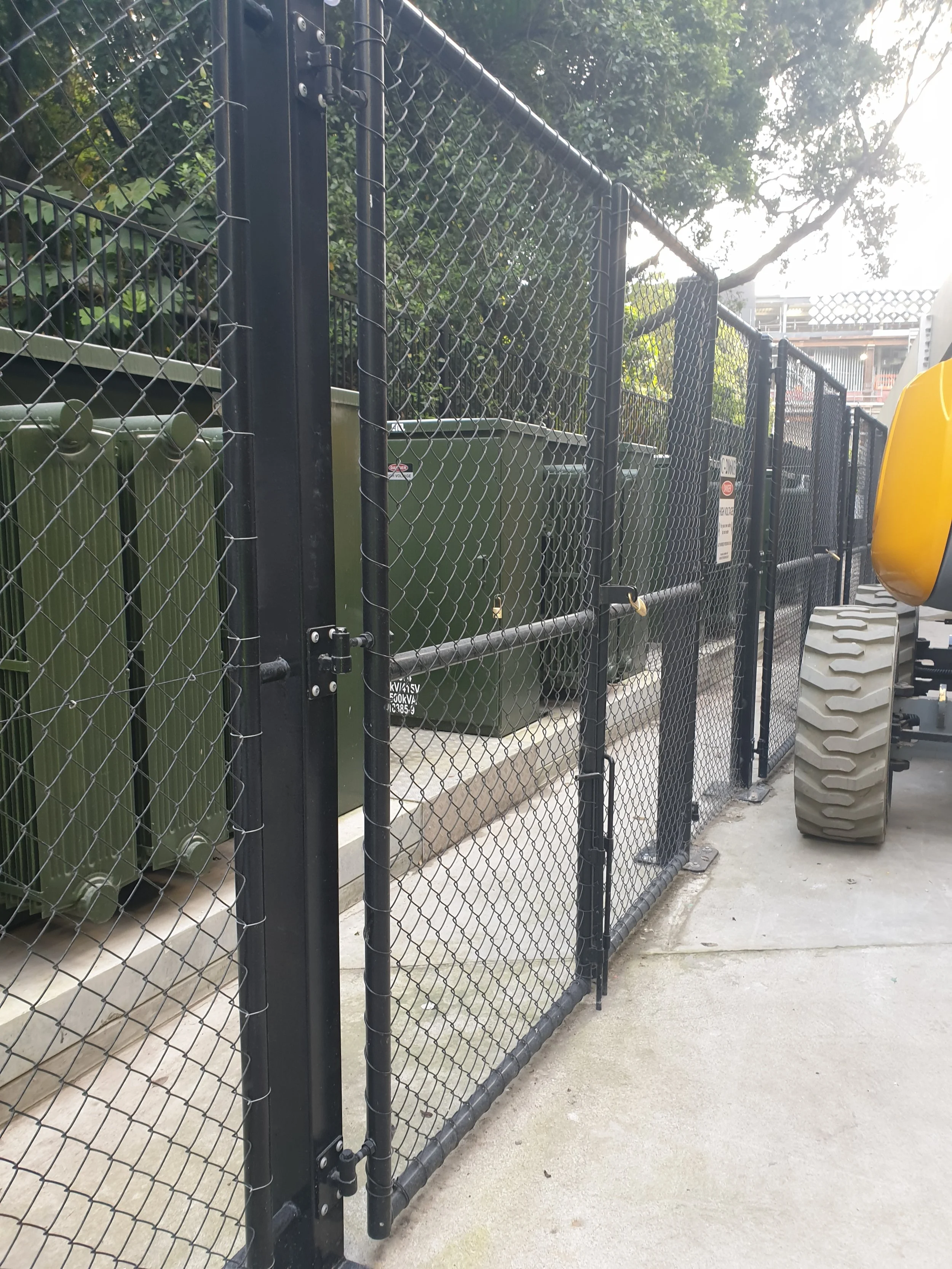 A black chain-link fence with a gate, behind which are green electrical utility boxes, and a gray concrete sidewalk in front. There are trees and a building in the background.