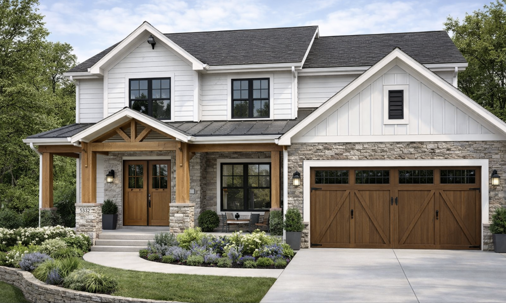 Front view of a modern two-story house with white siding, stone accents, wooden garage doors, and a covered porch with wooden posts. There are landscaped flower beds and greenery in the yard.