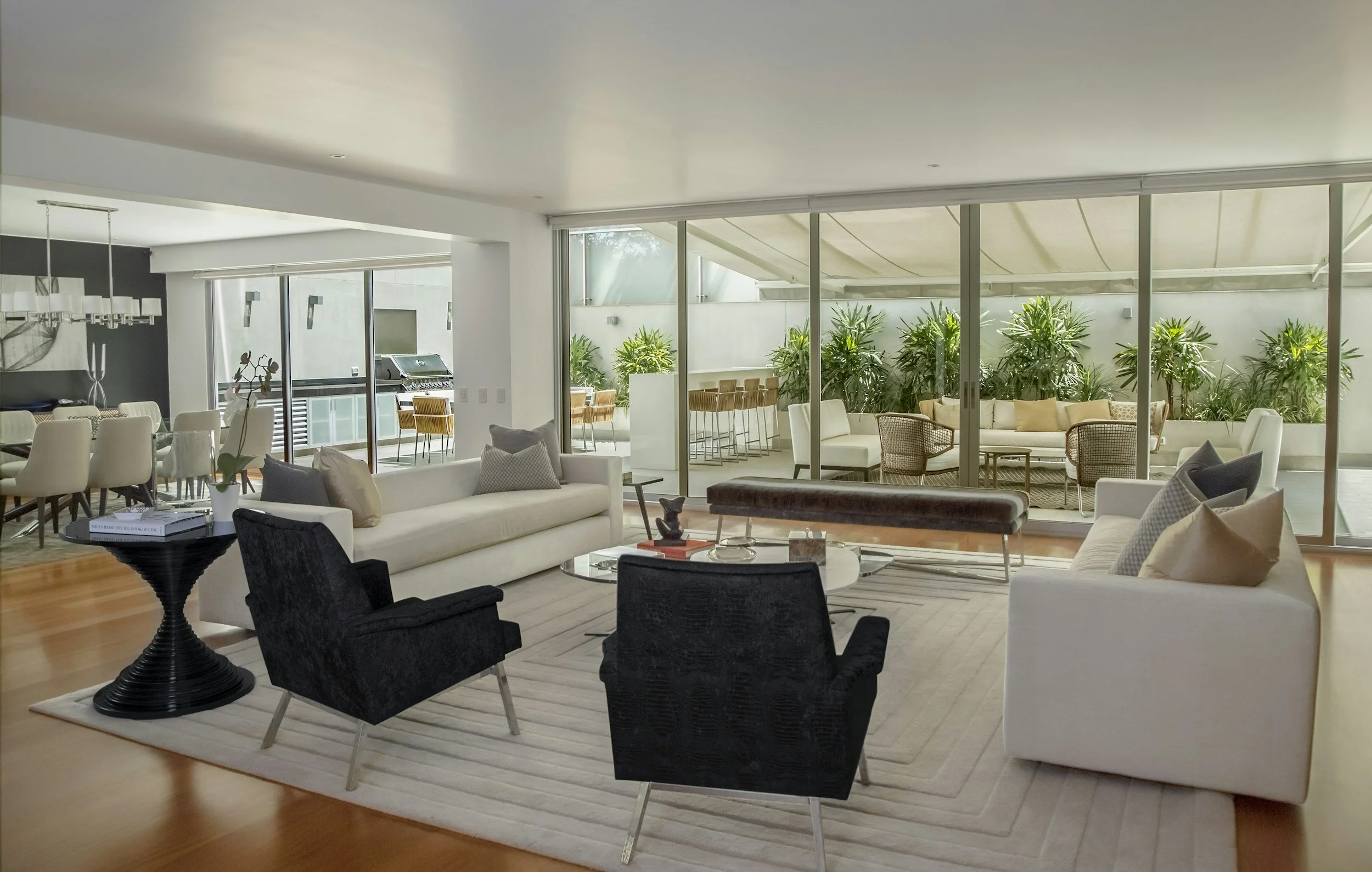 Modern living room with white sofas, black and white armchairs, glass coffee table, and floor-to-ceiling windows showing an outdoor patio with plants and seating area.