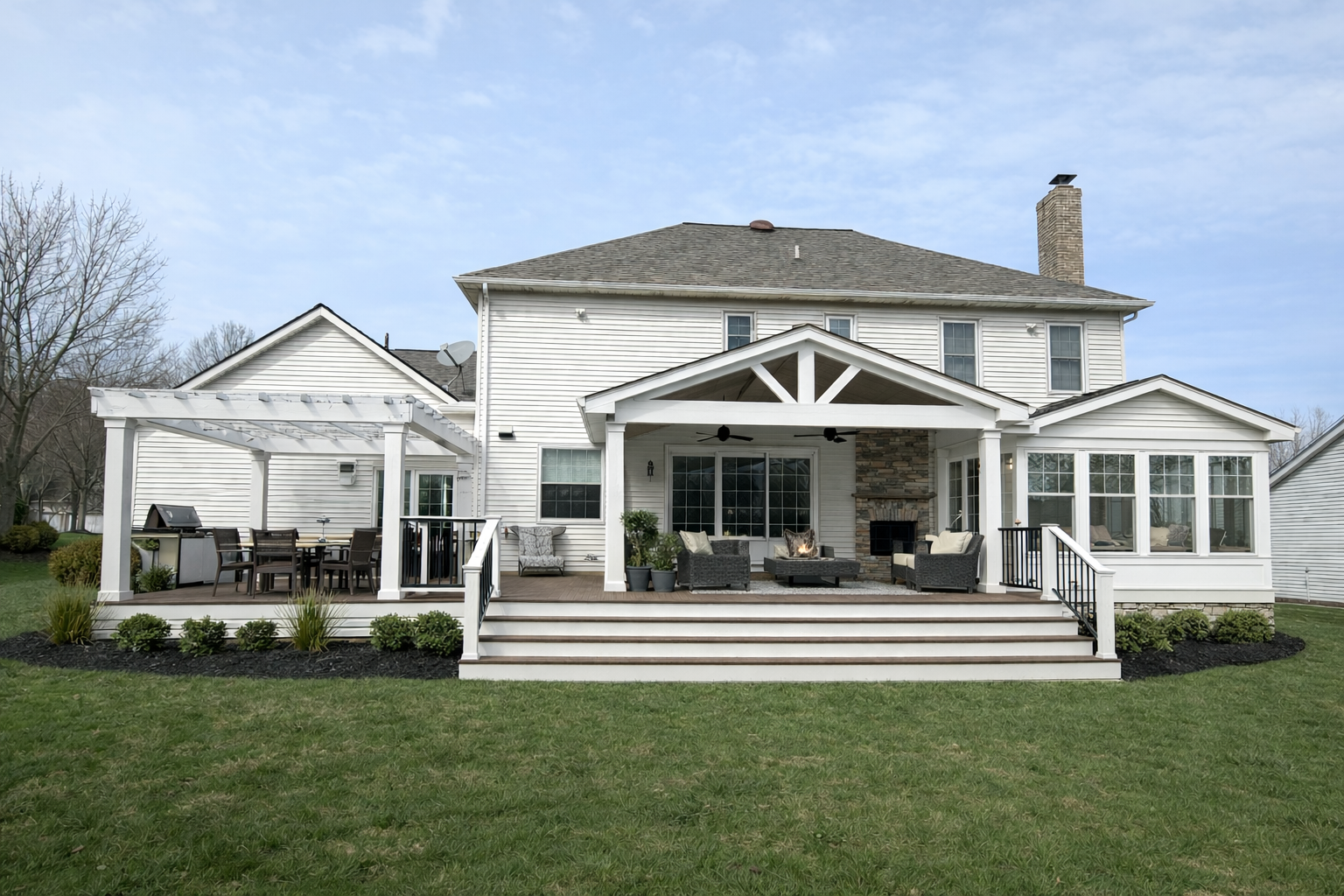 Large white two-story house with a spacious backyard patio featuring outdoor furniture, a fireplace, and a dining area. The patio has multiple levels with steps leading down to a grassy lawn.