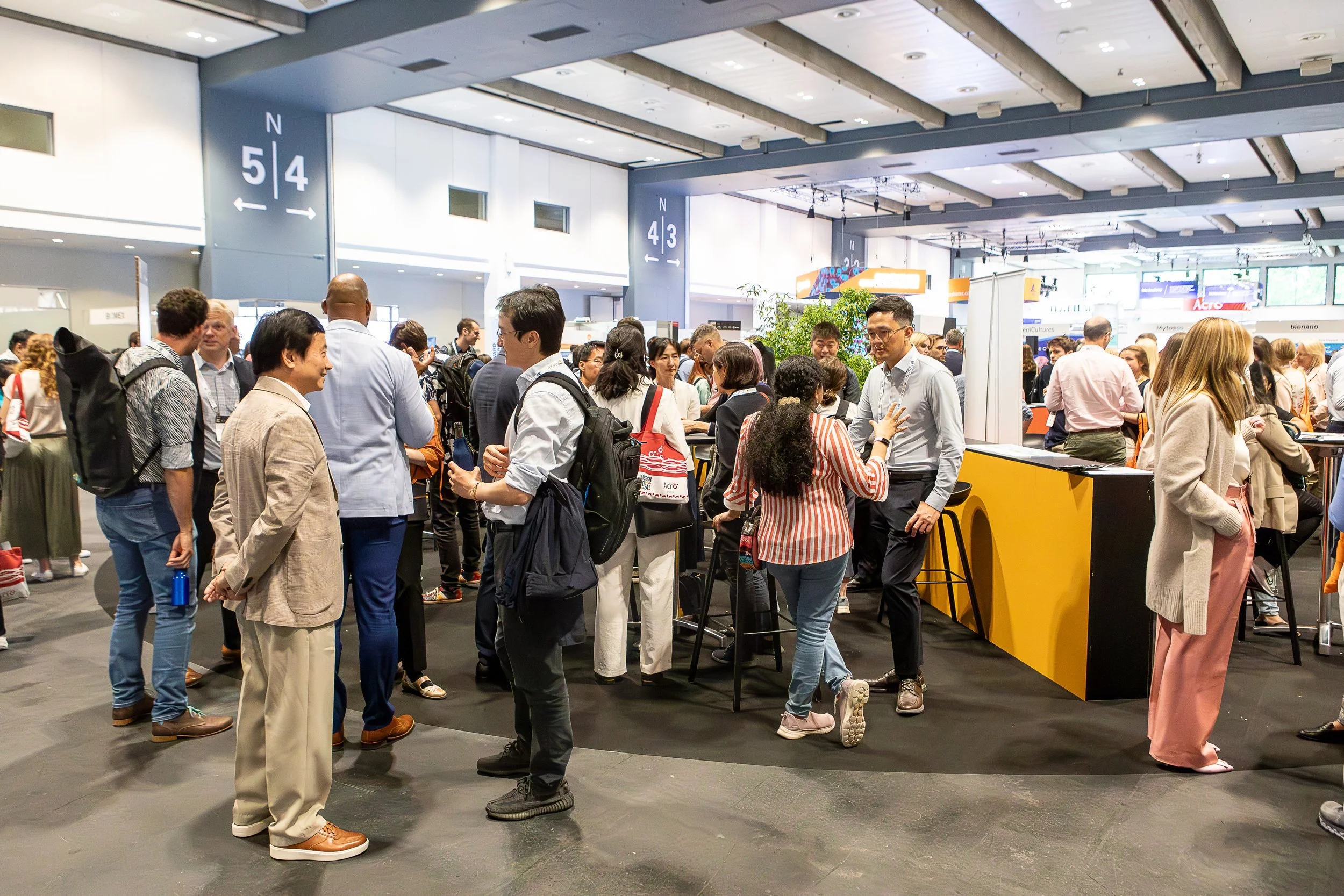 People gather at a busy convention or trade show with registration booths, signs, and informational displays in a modern, well-lit conference center.