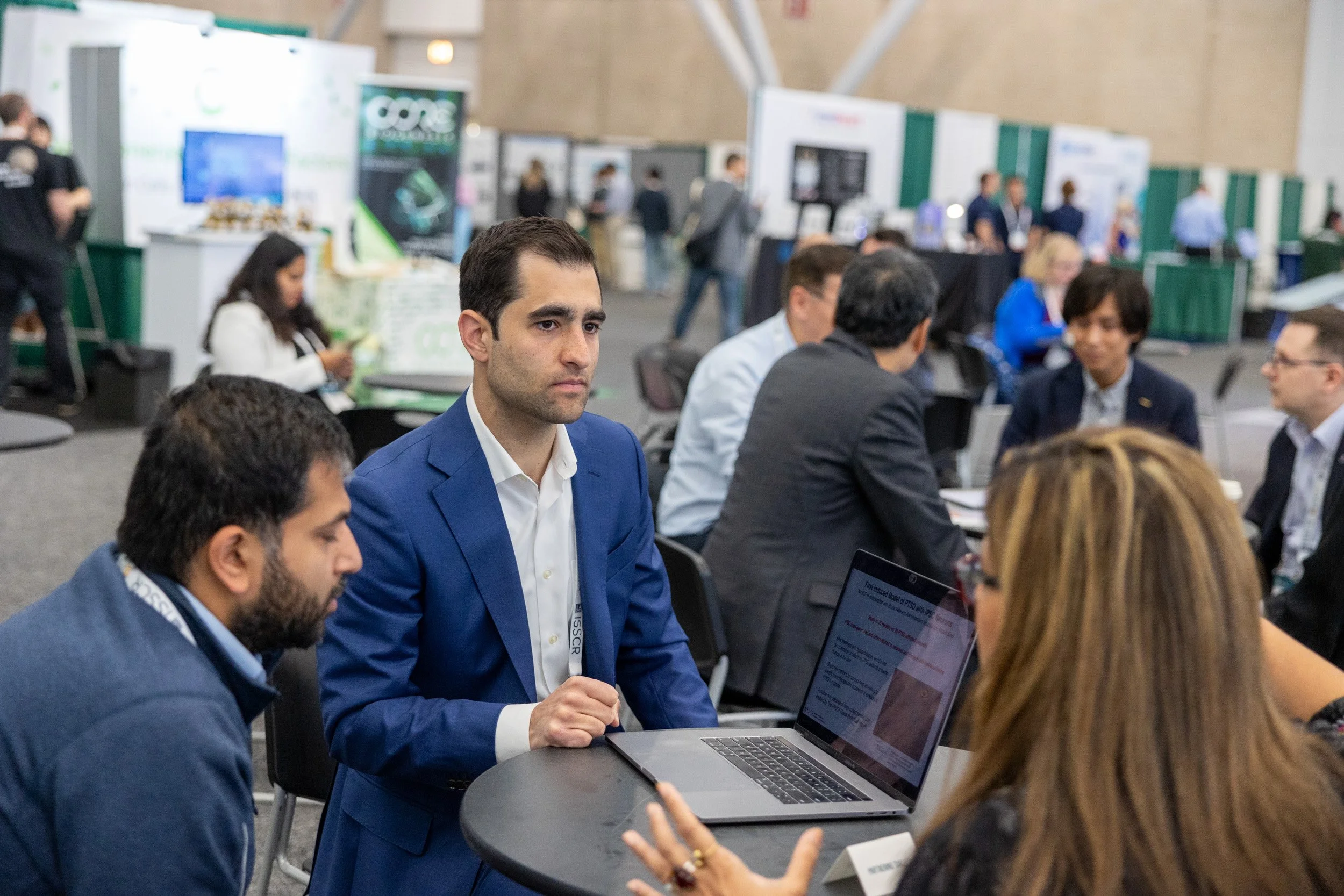 Business professionals at a conference or networking event, engaged in conversation at a table with laptops and informational materials.