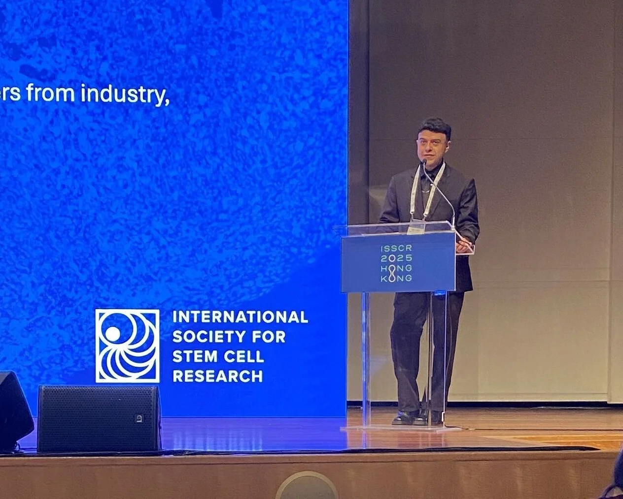 A man in a dark suit stands at a clear podium with a lanyard around his neck, speaking at the ISSCR 2025 Hong Kong conference, with a large blue presentation screen behind him displaying the logo of the International Society for Stem Cell Research.